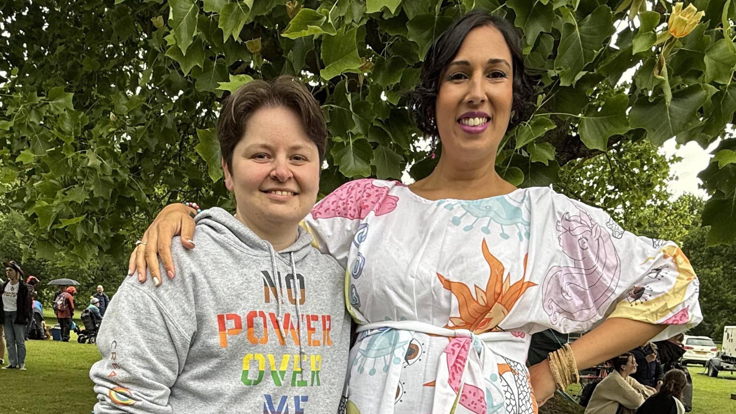 Riley Millar is stood on the left wearing a grey hoodie. AJ Singh on the right is standing with their arm over Riley Millar, and is wearing a white patterned dress. They are both looking at the camera smiling.