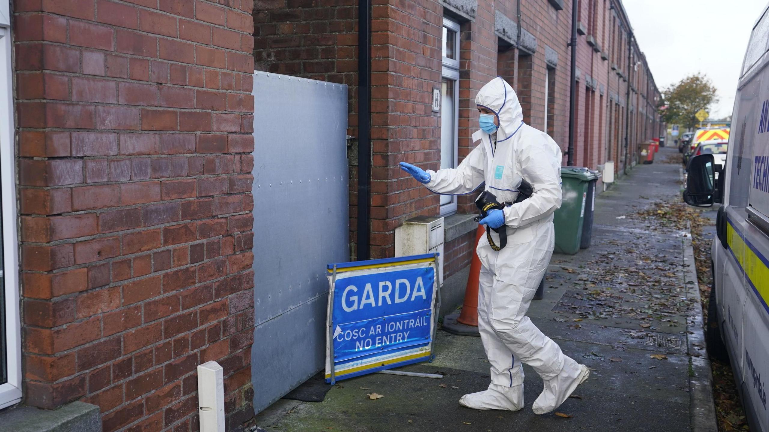 A forensic officer dressed in a full white boiler suit carries a large camera towards a sheeted steel gate, marking the entrance to an alleyway. A small blue pop-up sign sits on the ground next to him which reads "Garda". Below that writing in Irish reads "Cosc ar iontrail" followed by the English translation of "no entry". A Garda van is parked behind the forensic officer and in the background are some wheelie bins and red bricked terraced houses.