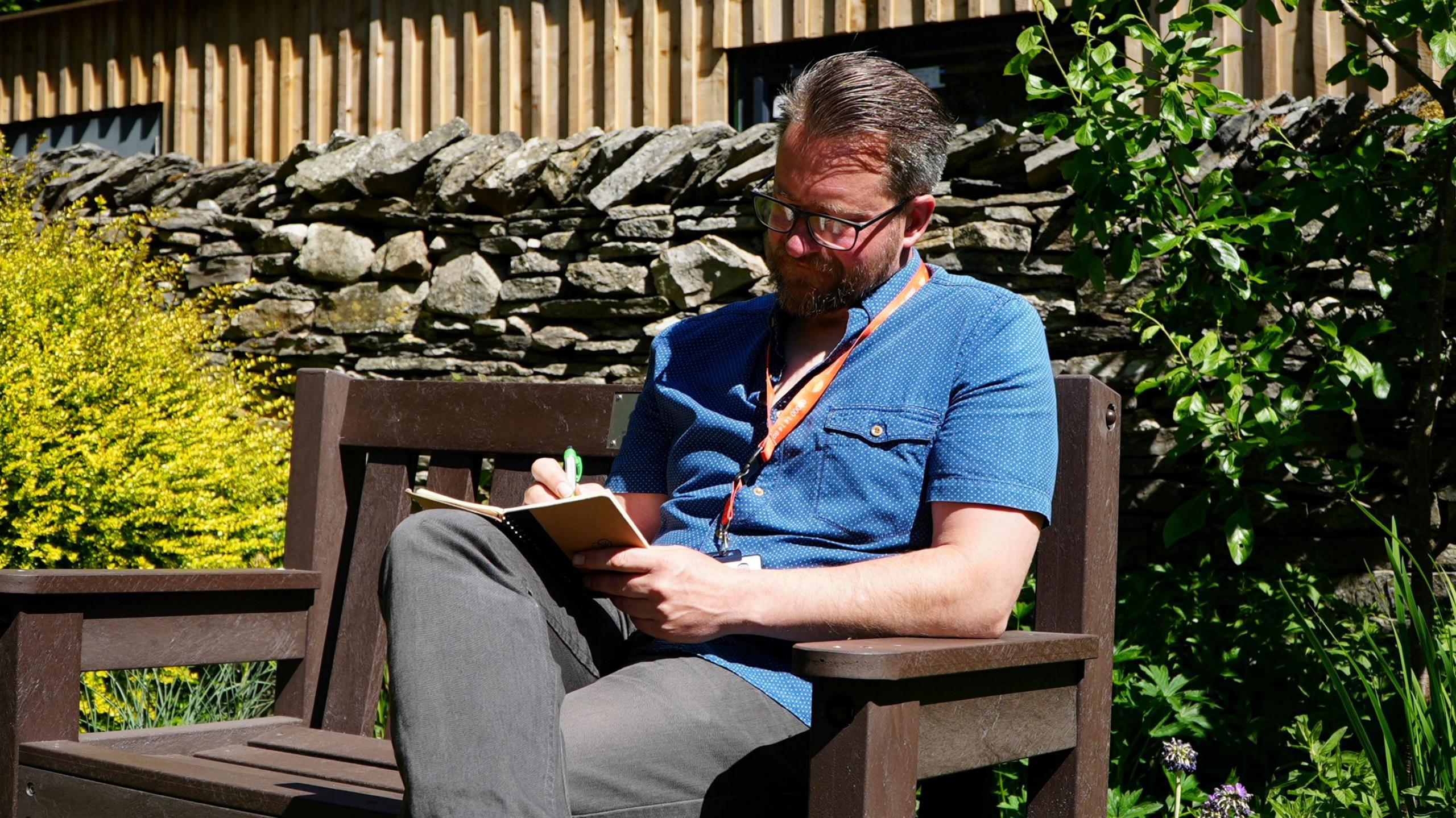Jimi Wilson wearing a blue short-sleeved denim shirt sitting on a park bench and writing in a notepad. He has an orange lanyard around his neck and is wearing black framed square glasses.