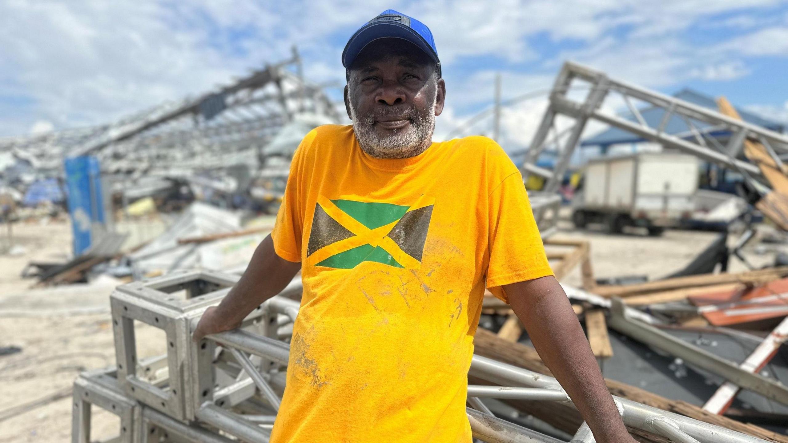 A man wearing a bright yellow shirt with the Jamaican flag on it is seen leaning on metal debris. Everything behind him is destroyed