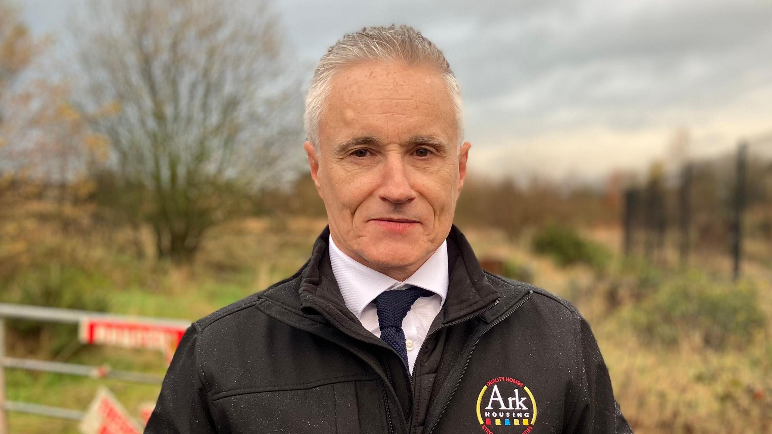 Jim McShane with short white hair stares at the camera as he wears a black "Ark Housing" branded coat, a white shirt and a navy tie. In the background is grass, trees and a silver metal gate.