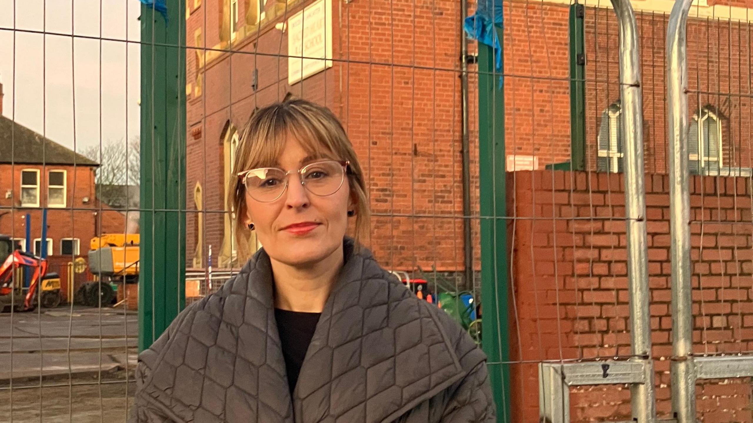 Simone Beach is standing in front of the red-brick two-storey brick building. She has blonde hair and is wearing glasses and a grey coat.