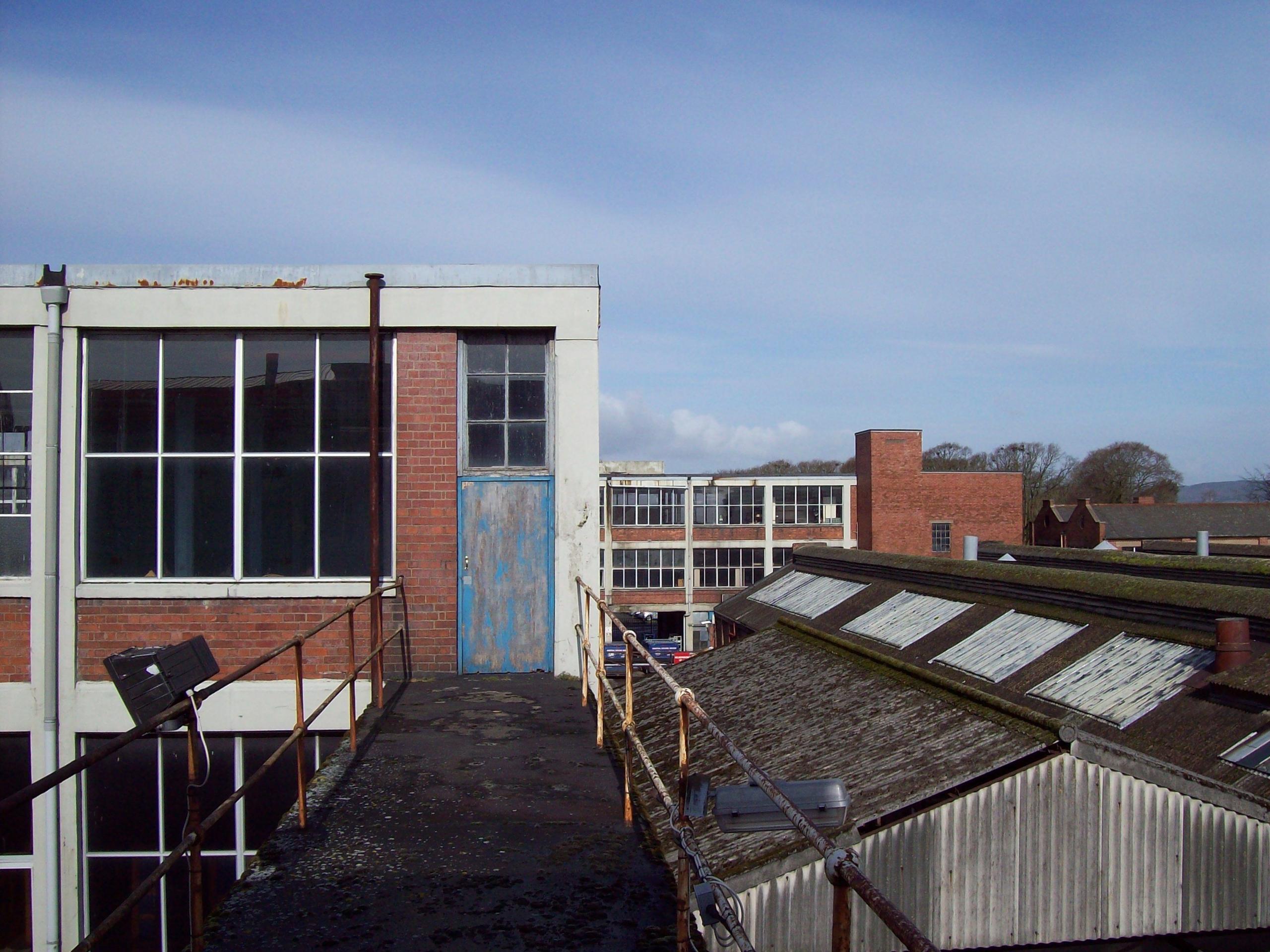 A rooftop view of a factory with metal railings and corrugated iron roofs against a blue sky