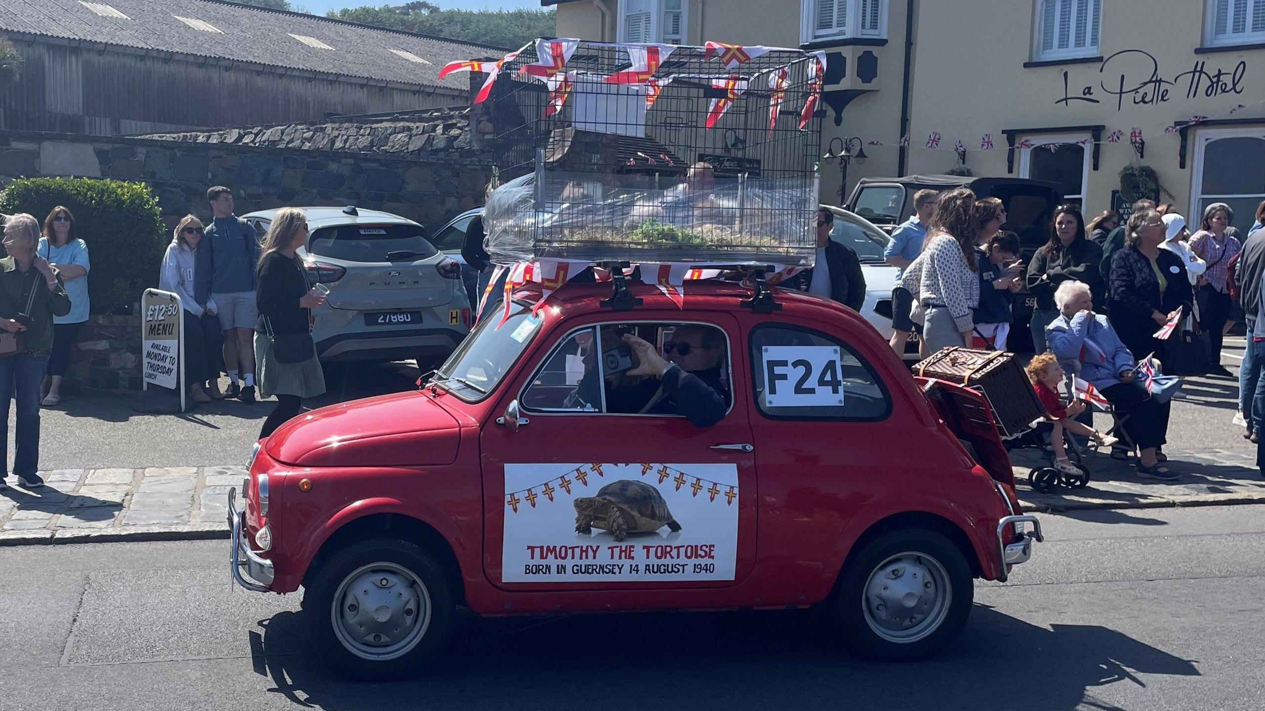 A red classic fiat 500 taking part in the cavalcade. A large cage is on top of the car. Inside you can see the tortoises shell and some staw.