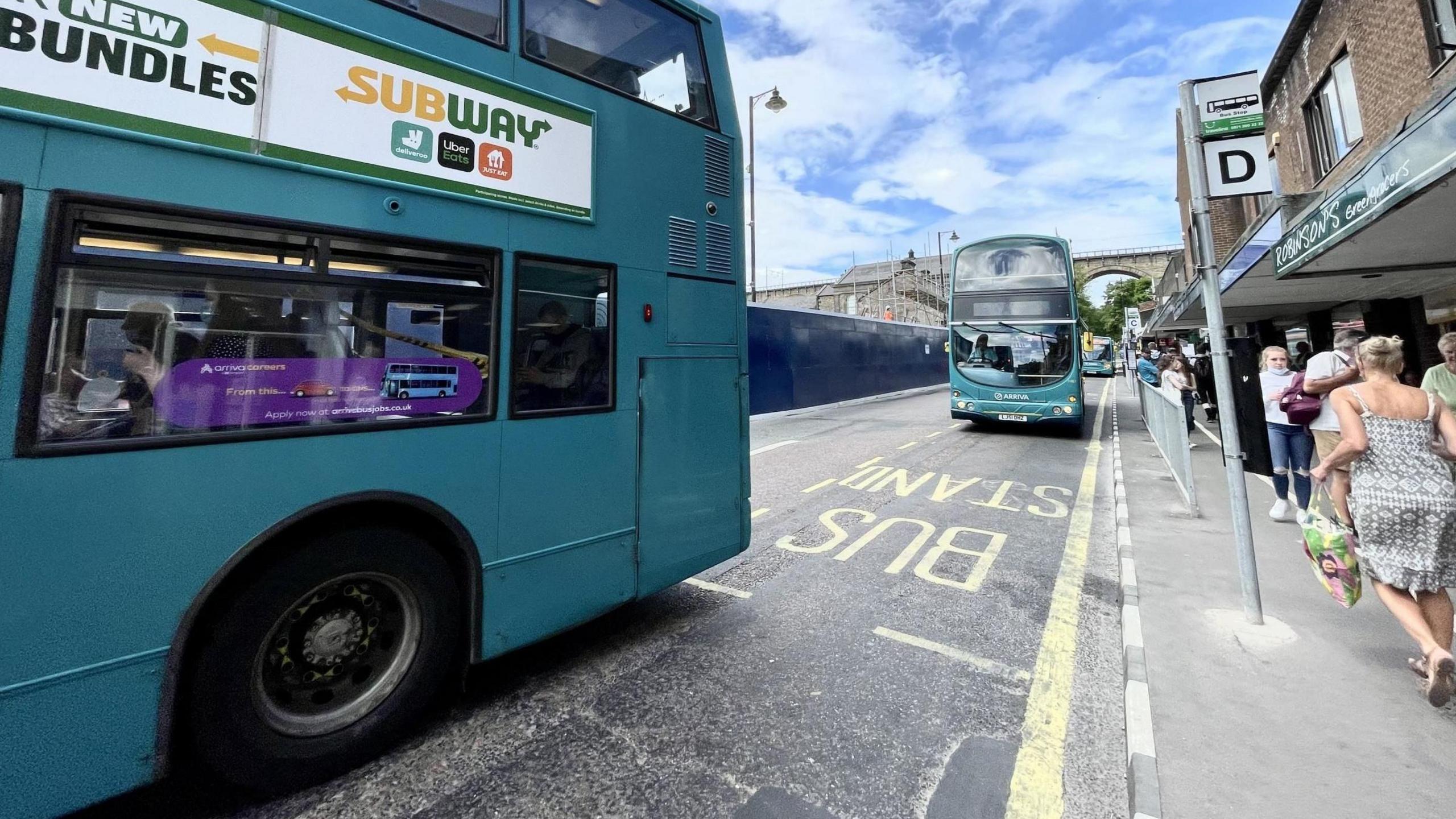 Two Arriva buses on a road. The vehicles are painted blue. Several people are waiting at a bus stop on the pavement.