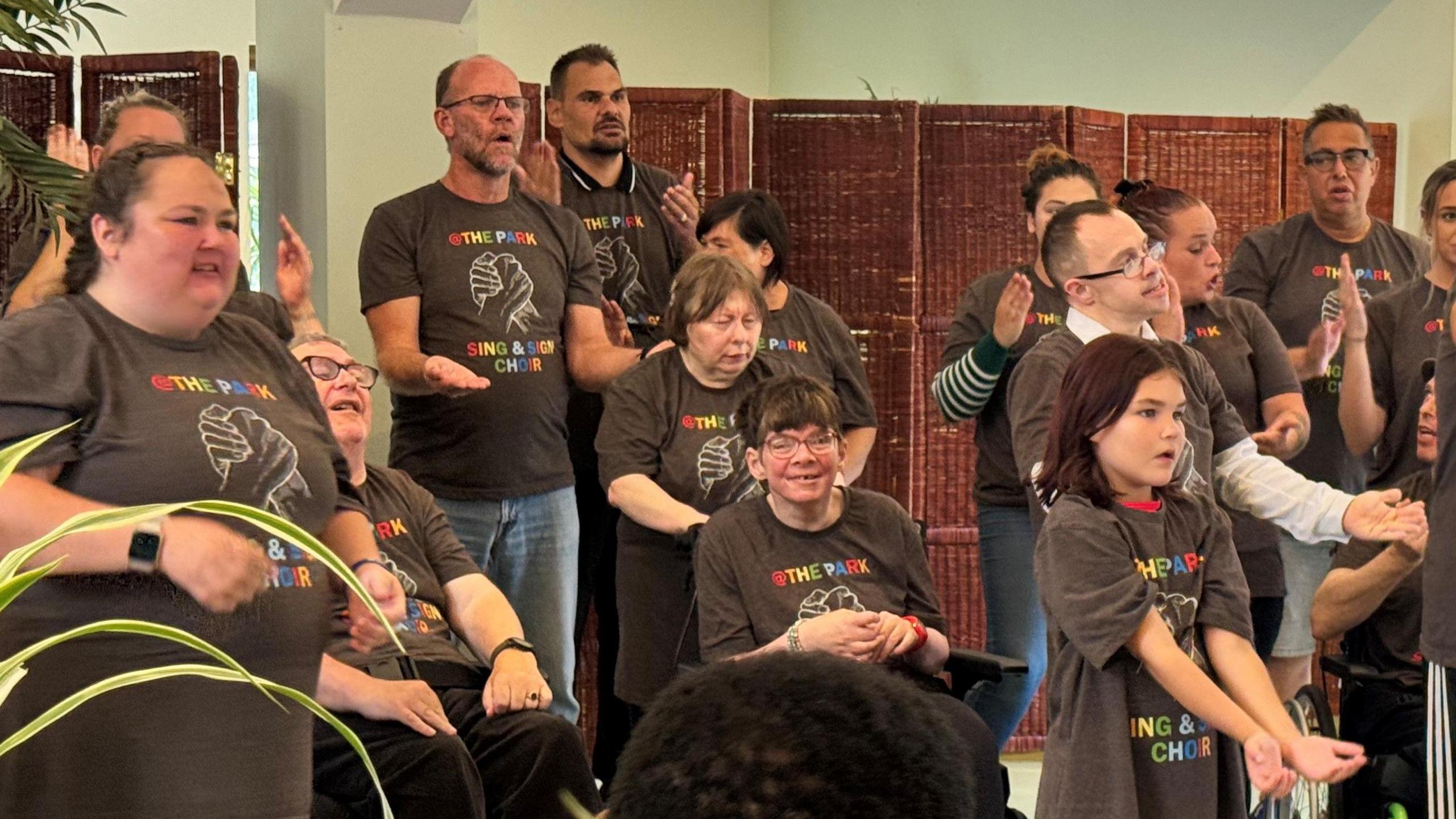 Close up of some of the members of the At The Park Sign and Sign Choir. On the far left is Penny, a choir member. She has long dark hair which is tied back. She is among a group of choir members who are signing and singing.