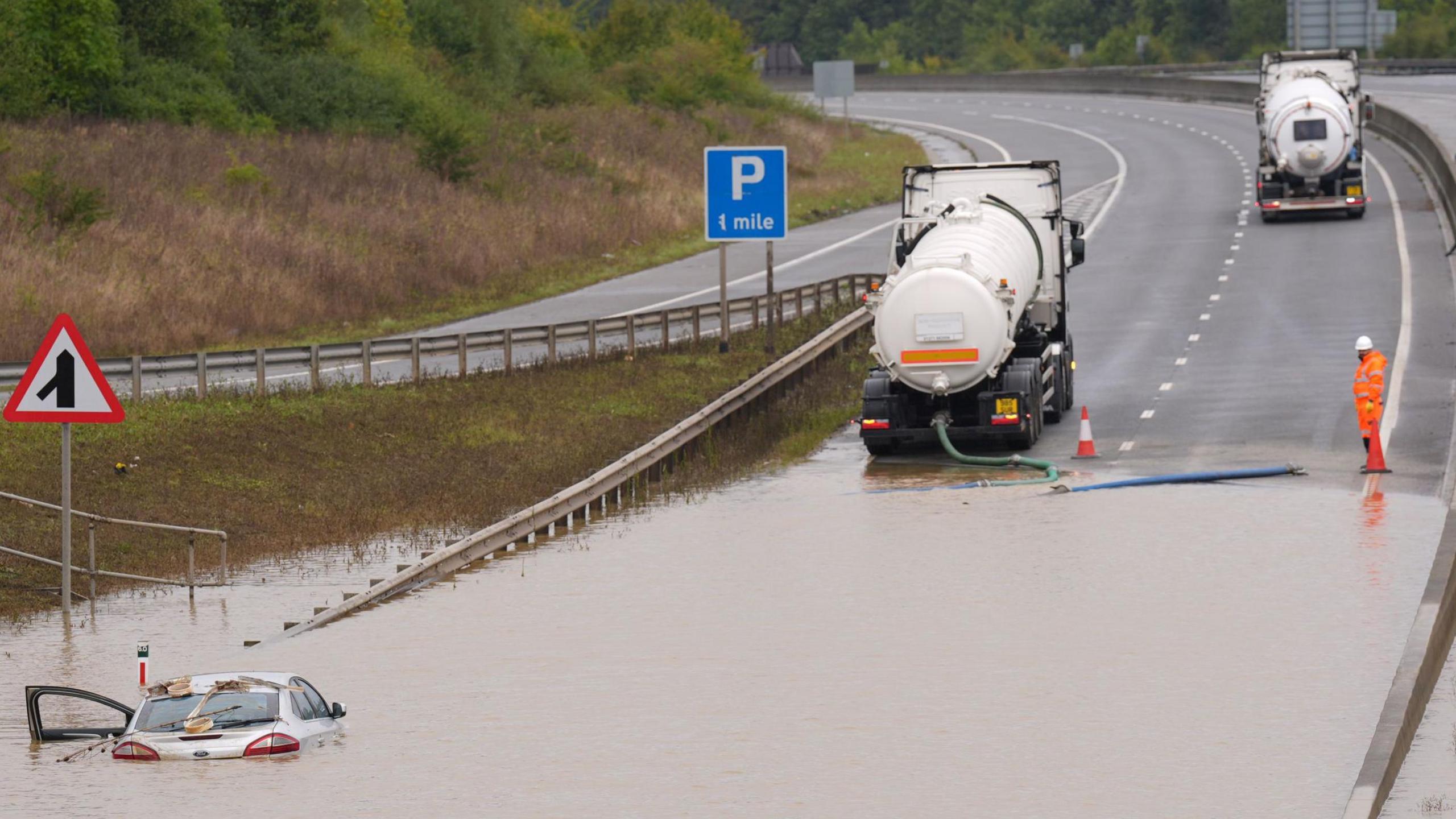 A tanker parked up next to a large body of water, which covers a dual carriageway. A green pipe can be seen coming out of the back of the tanker and leading into the water. A silver car is partially submerged in the water and has been left with its front passenger door open. Another tanker can be seen in the distance, apparently reversing towards the body of water. In the distance is a slip road leading on to the dual carriageway and a steep grass verge.