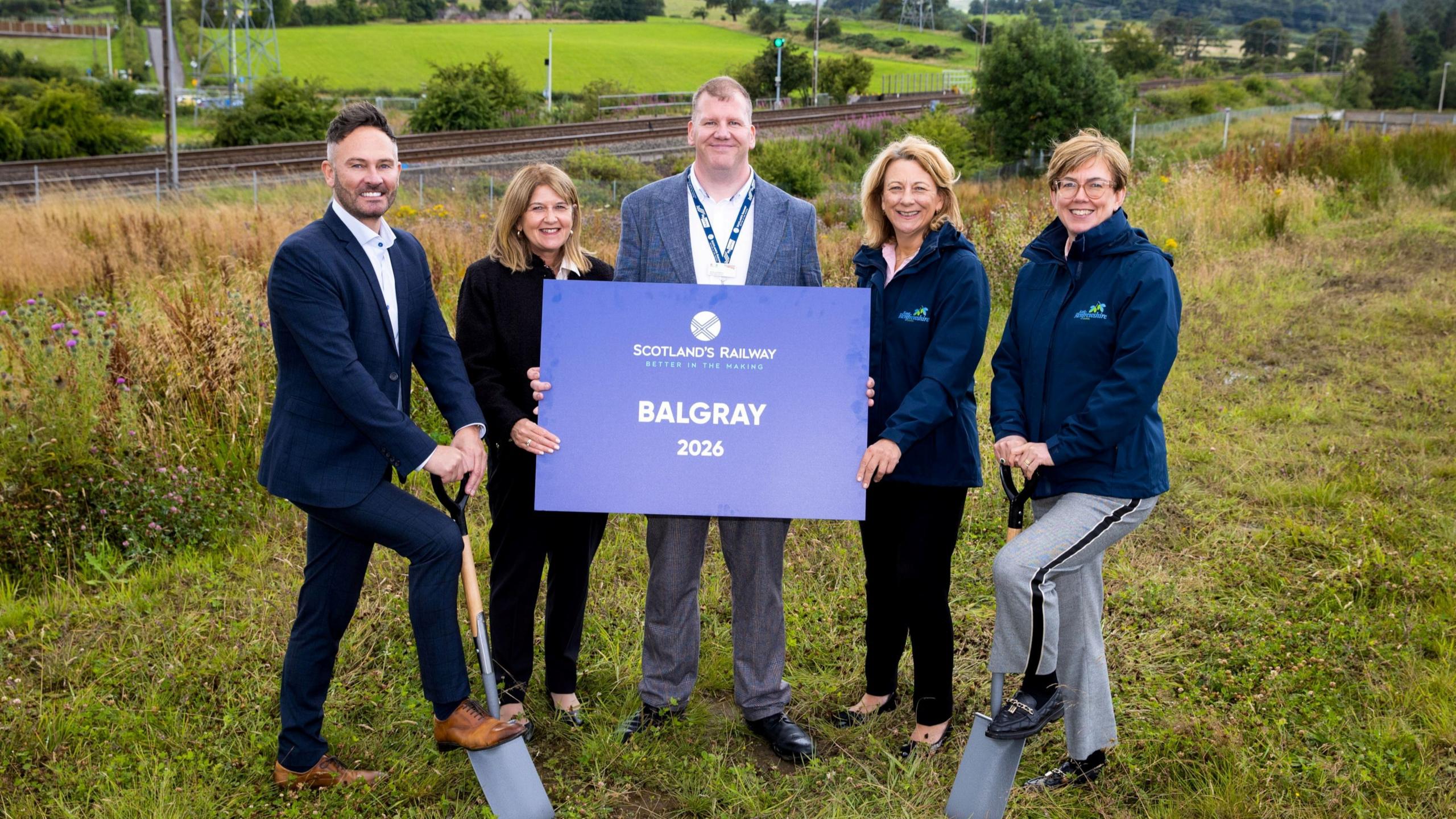 Five people - two men and three women - stand on a grassy area. The man and woman at either side of the row have shovels digging into the ground, while the man in the middle, wearing a grey suit, is holding a sign saying BALGRAY 2026.
All of the group are smiling towards the camera.