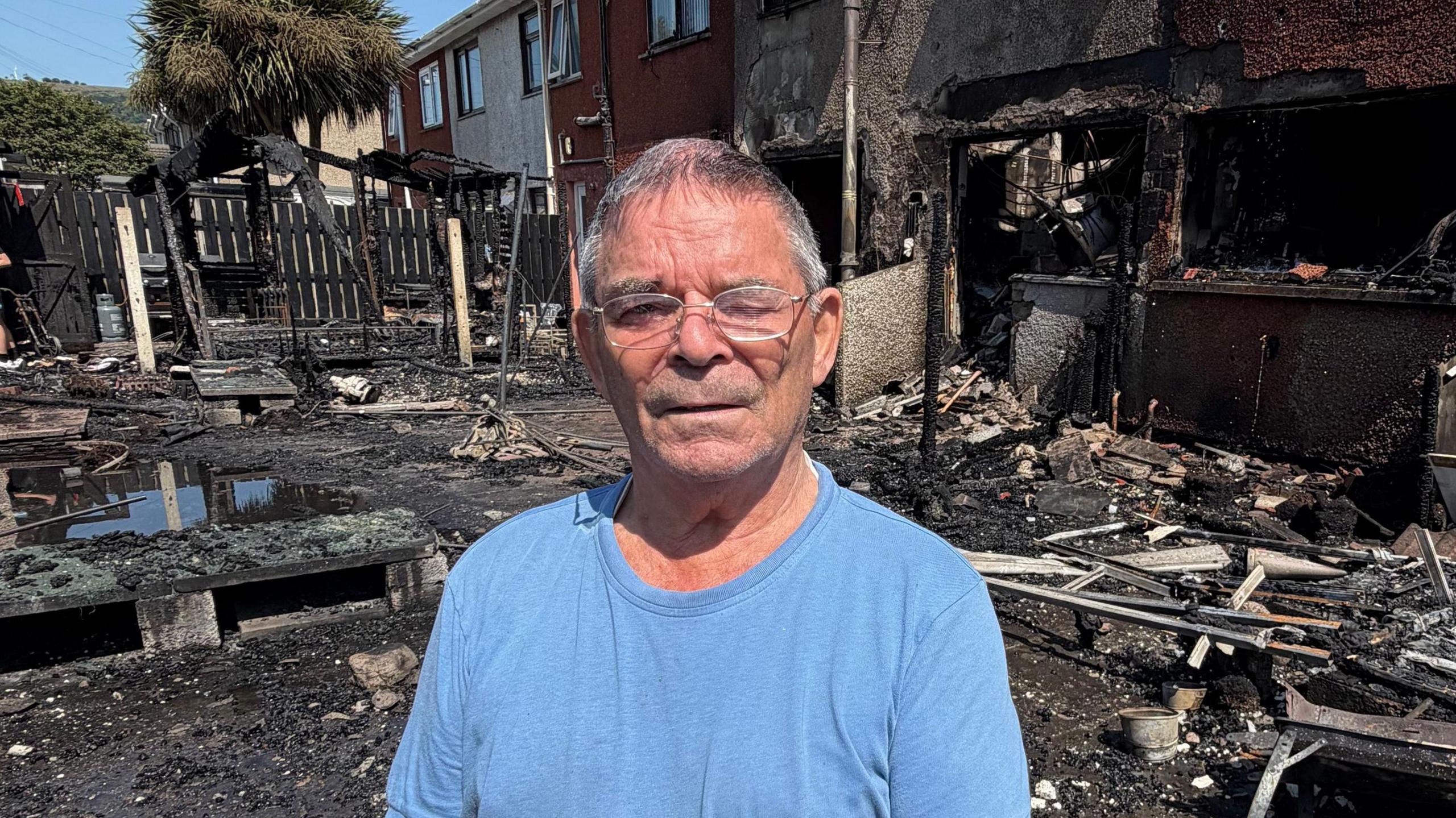 Davy Haighton, a man with short grey hair and silver-framed glasses stands in front of the wreckage of two terraced houses which have been gutted by fire. He is wearing a light blue crew-neck t-shirt