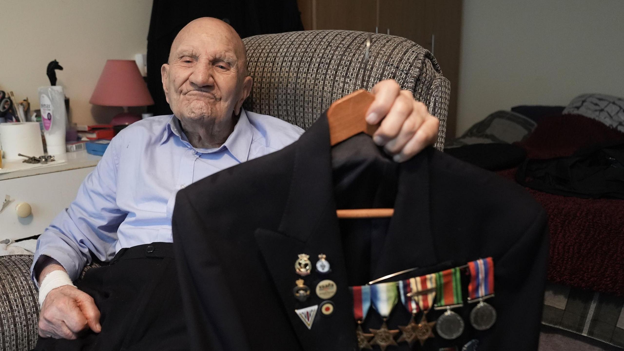 A man sitting on a chair, wearing a blue shirt and black trousers and looking towards the camera. His left hand is holding up to the camera a black jacket with medals on it.