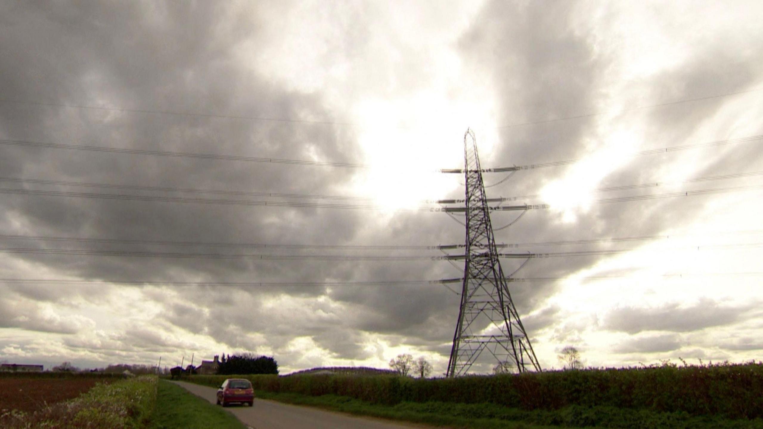 An image of a pylon in front of a cloudy sky with power lines running to the left and right. There is a road in the foreground with a red car travelling along it.