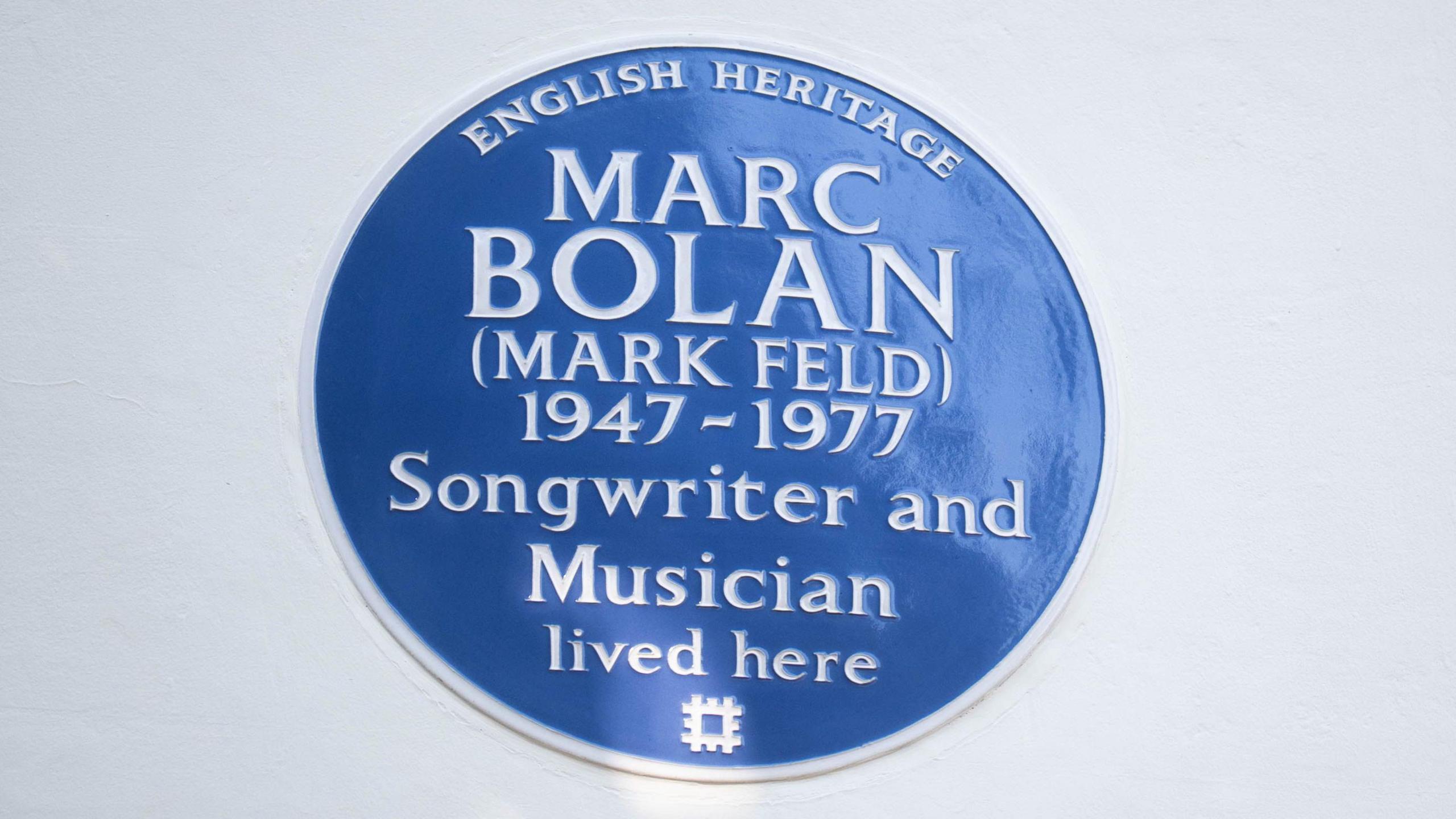 A close up of a blue English Heritage plaque on a white wall. 