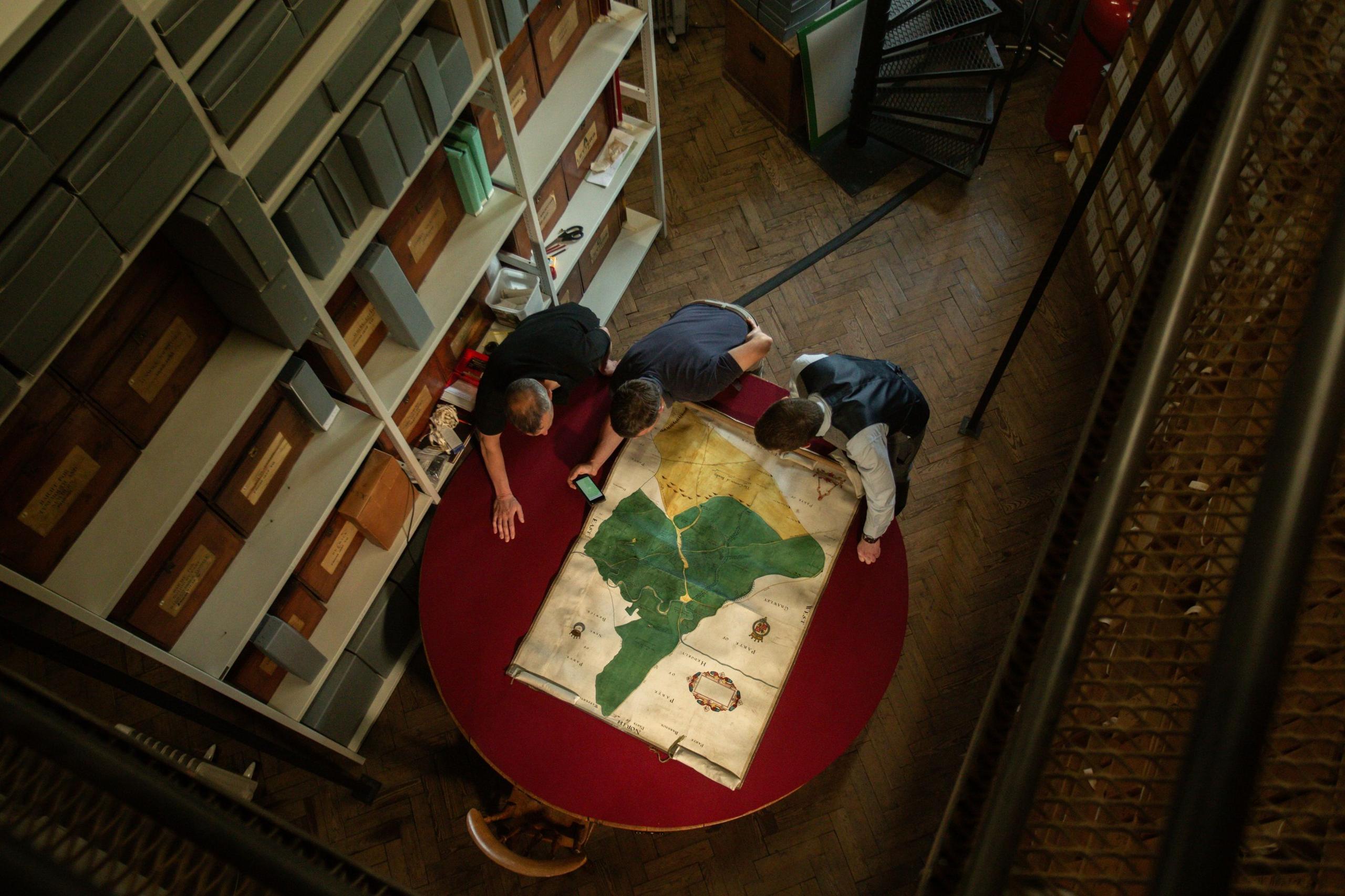 Three men lean over a map that is spread out on a red, wooden circular table in a dimly lit room, which also contains large, tall shelves with various boxes on.