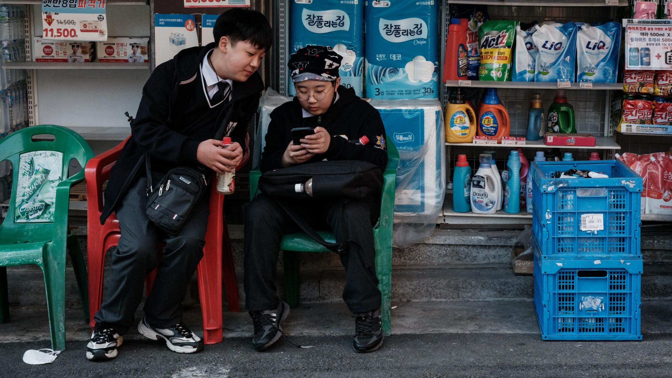 A South Korean schoolboy, wearing a school uniform and glasses, scrolls on his phone while sitting in a plastic chair outside a convenience store. Another boy in the same uniform leans over from his own chair to watch the screen.