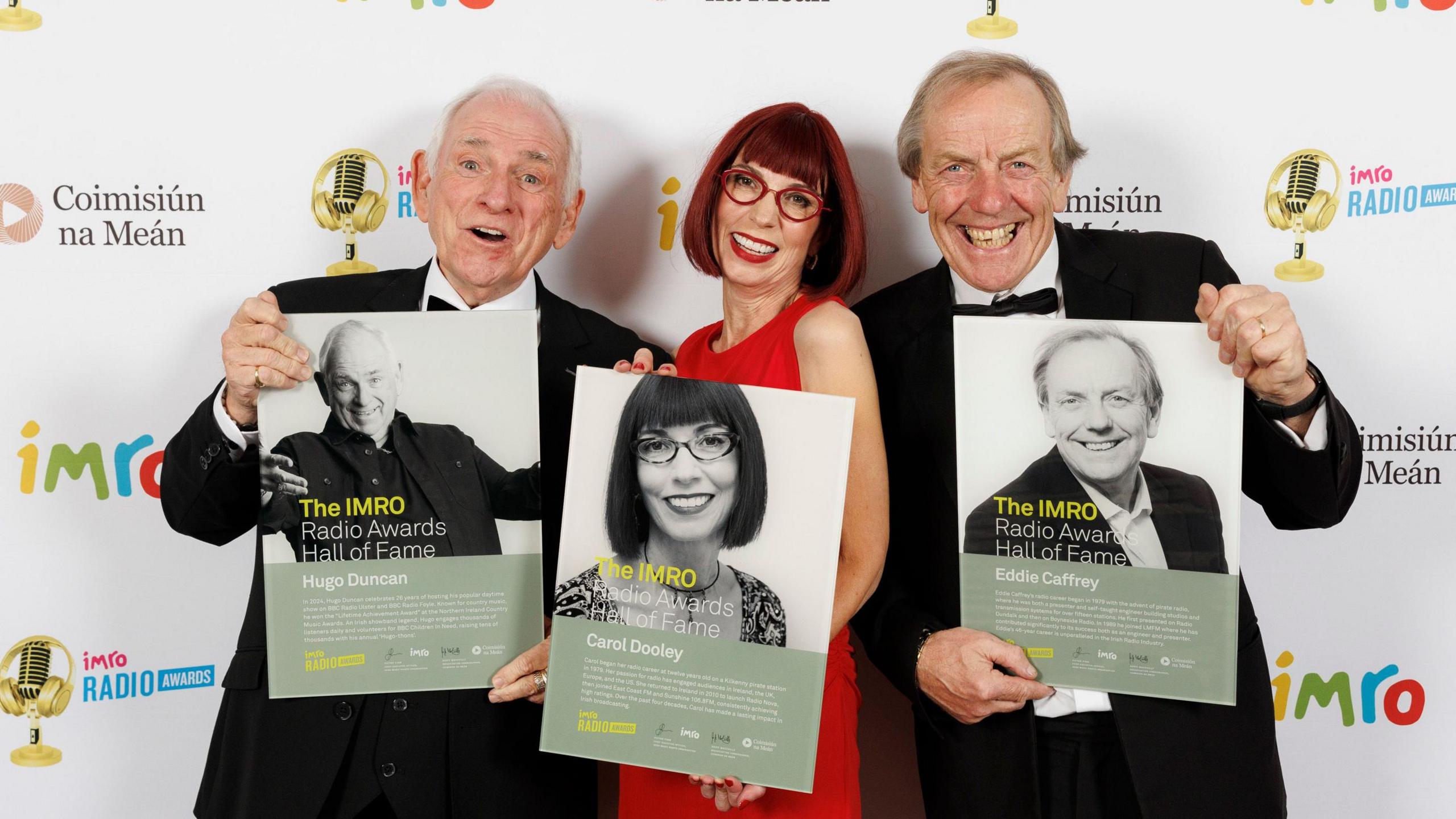 Hugo Duncan at the IMRO awards wearing a suit and holding his Hall of Fame Plaque, he is standing beside Carol Dooley (dressed in red with red glasses and dress) and Eddie Caffrey in a black suit.