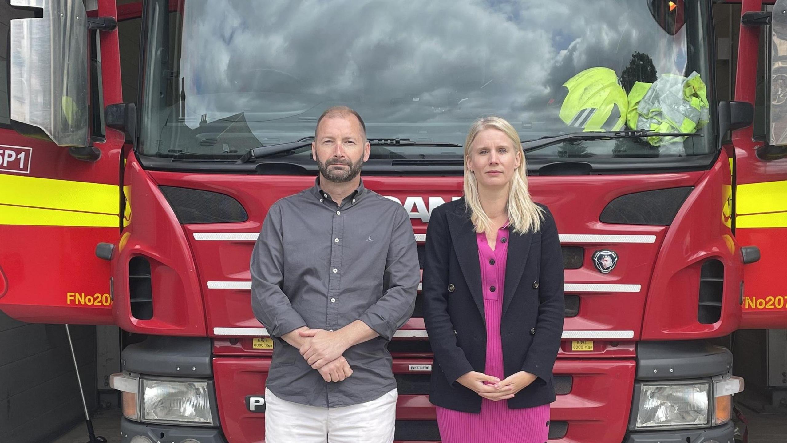 A man and a woman stand in front of a fire engine at Banstead Fire Station. They have neutral facial expressions.