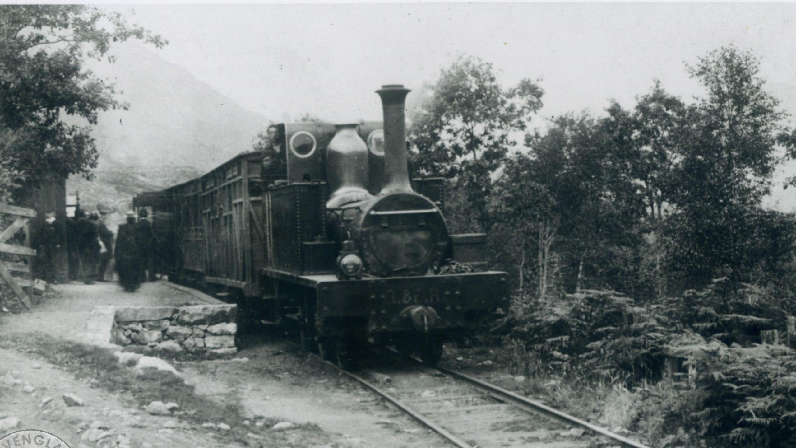 Black and white photograph, dating from 1875 of a steam train viewed from the front, with containers, rather than carriages behind it. It appears to be stationery on a platform on which a group of people are gathered. The surrounding are rural, with a mountain or large hill in the distant background.