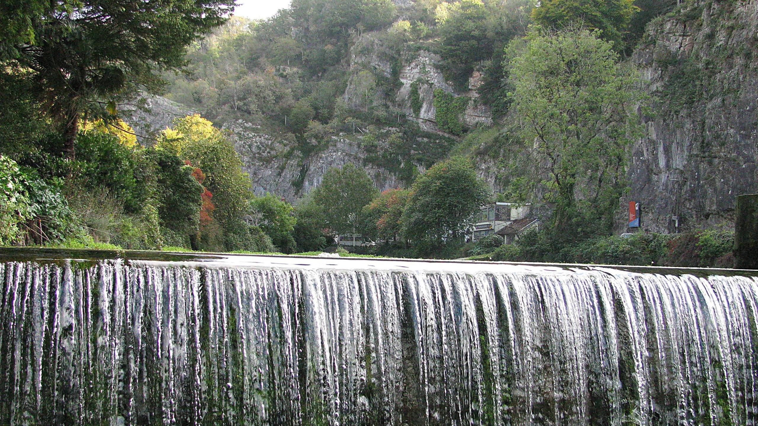 A wide waterfall spans the picture, and behind it is Cheddar Gorge, with high rocks and lots of green trees on the cliffs and river banks.