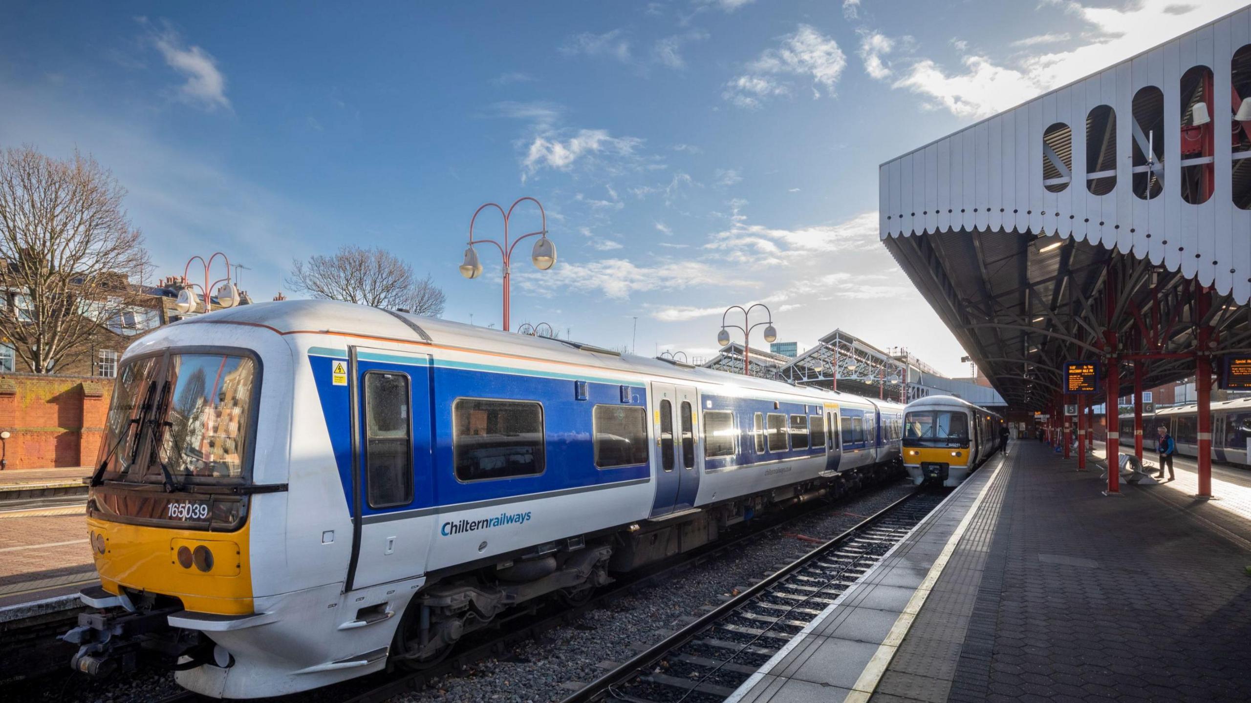 A Chiltern Railways train stopped at a train station tracks on a sunny day. Another one is seen approaching on the neighbouring track.