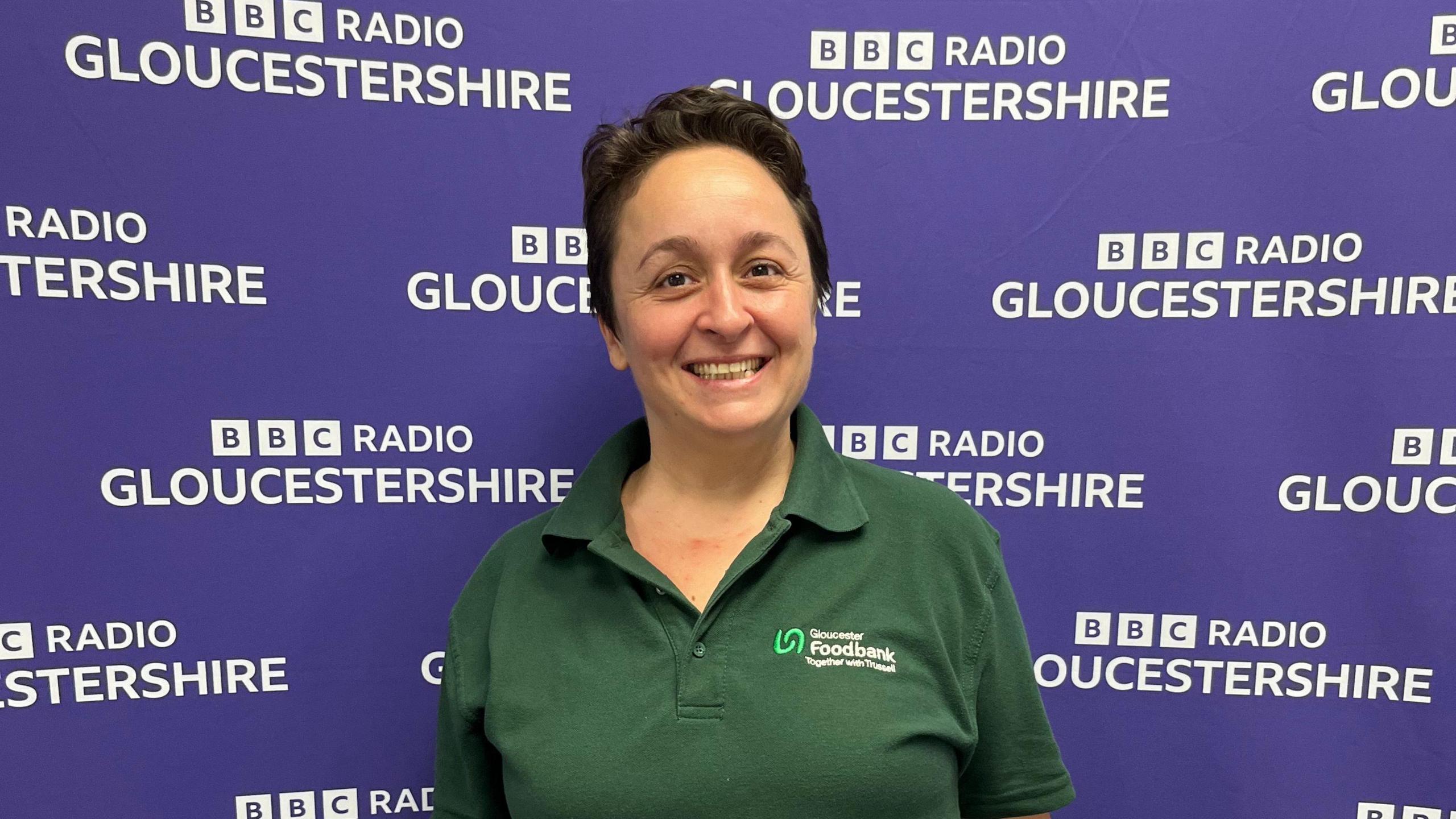 A smiling Vicky Ranford with her short dark brown hair wears a green polo shirt with the Gloucester Foodbank logo on the front. She is standing in from of the purple BBC Radio Gloucestershire branded back-drop.