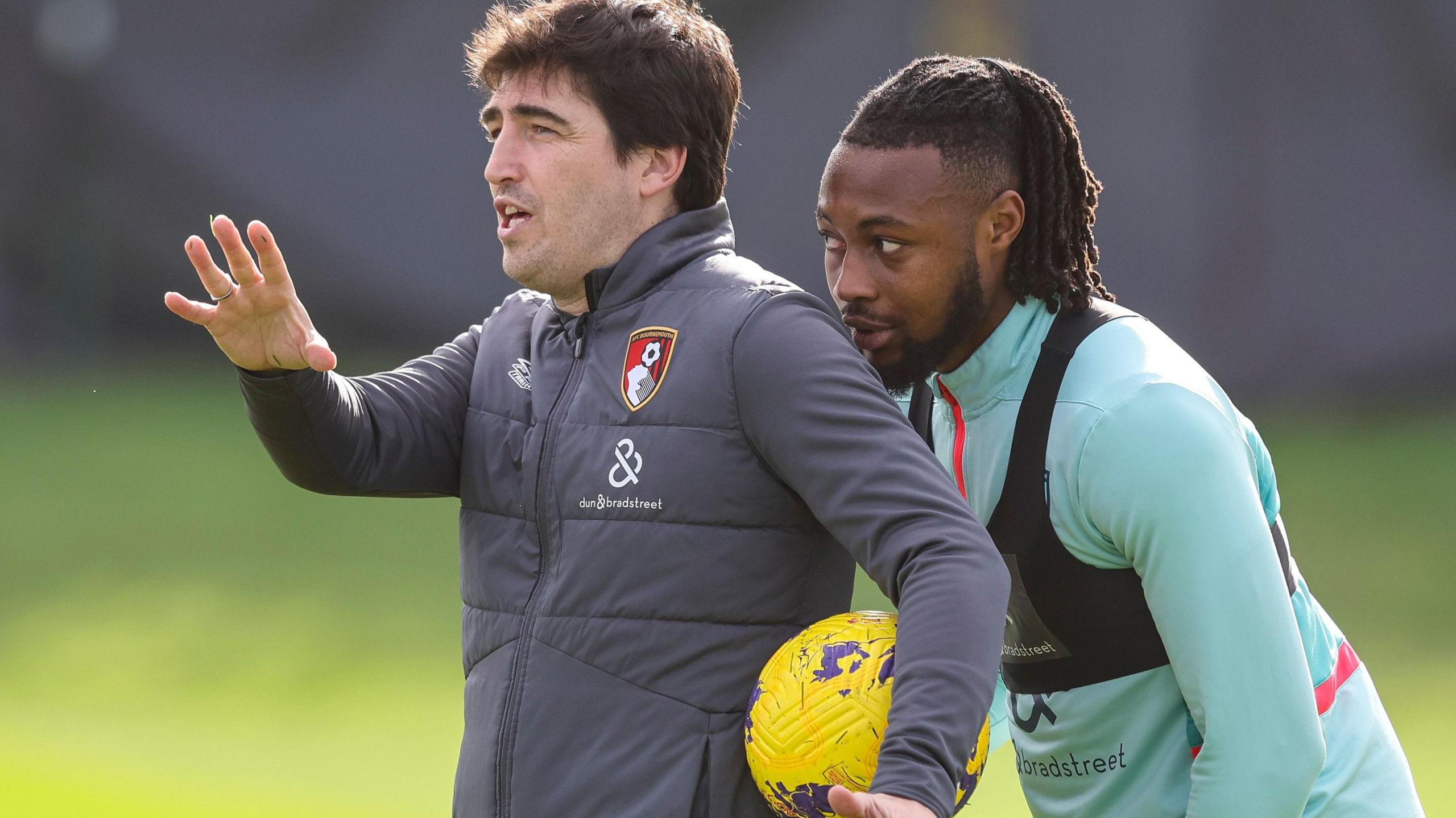 Antoine Semenyo leans in behind the left shoulder of Andoni Iraola, who is gesturing with his right hand while cradling a yellow football between his left arm and his torso