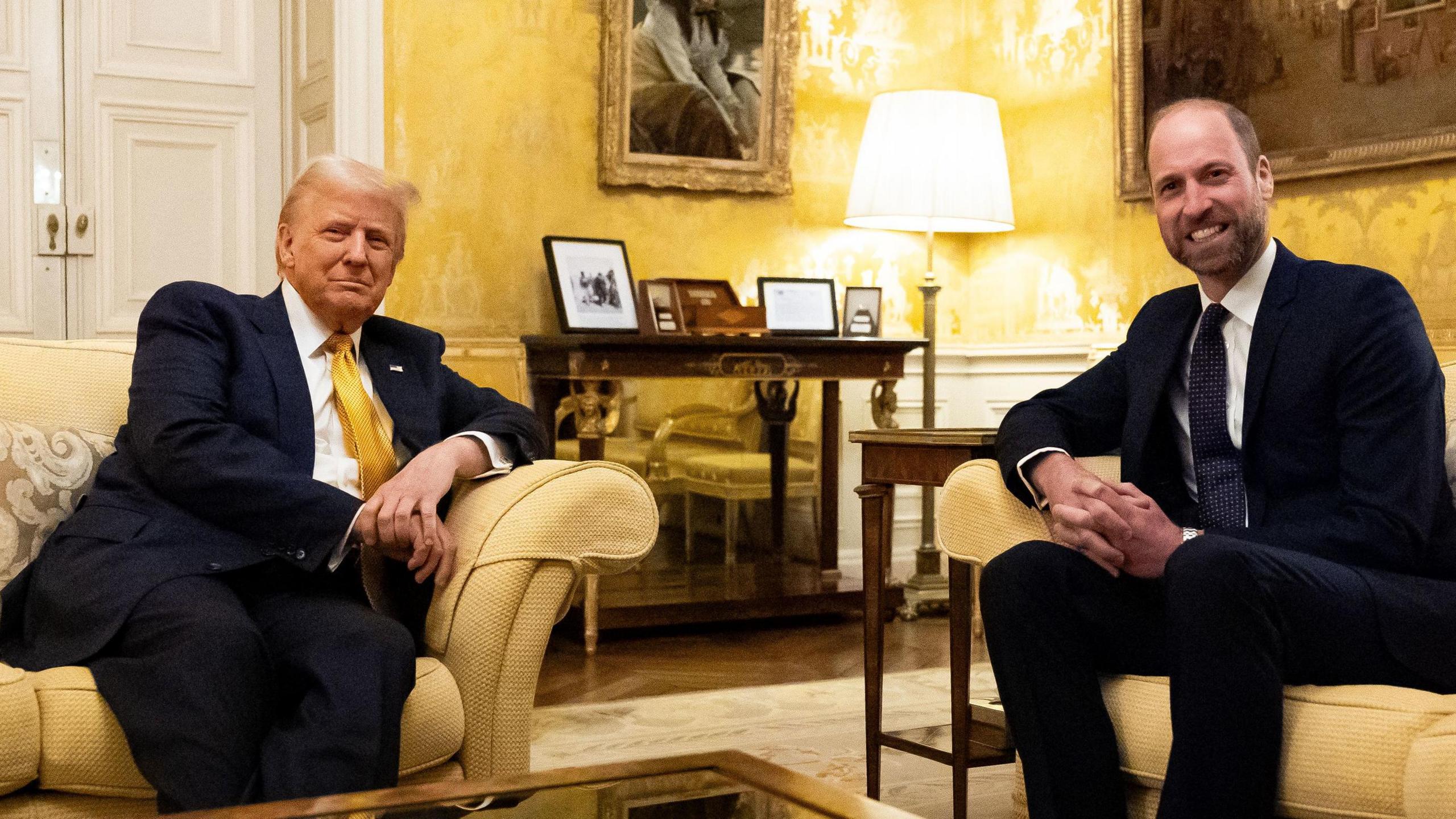 Donald Trump, who has blonde combed hair, sits on a yellow sofa and looks at the camera, as Prince William sits on a separate sofa and also looks at the camera