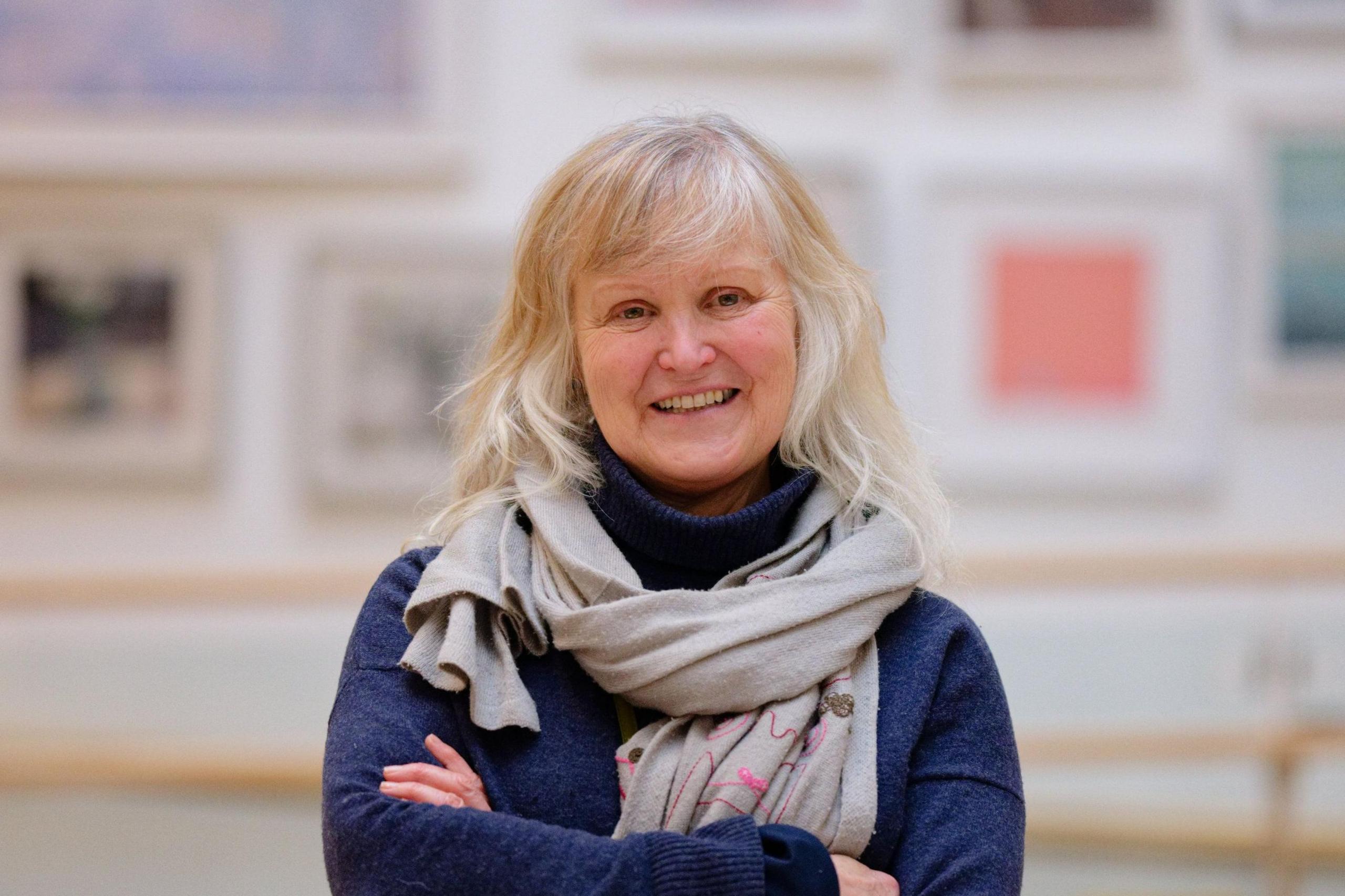 Andrea Perry stands with her arms crossed but is smiling. She wears a blue jumper and a grey, patterned, scarf