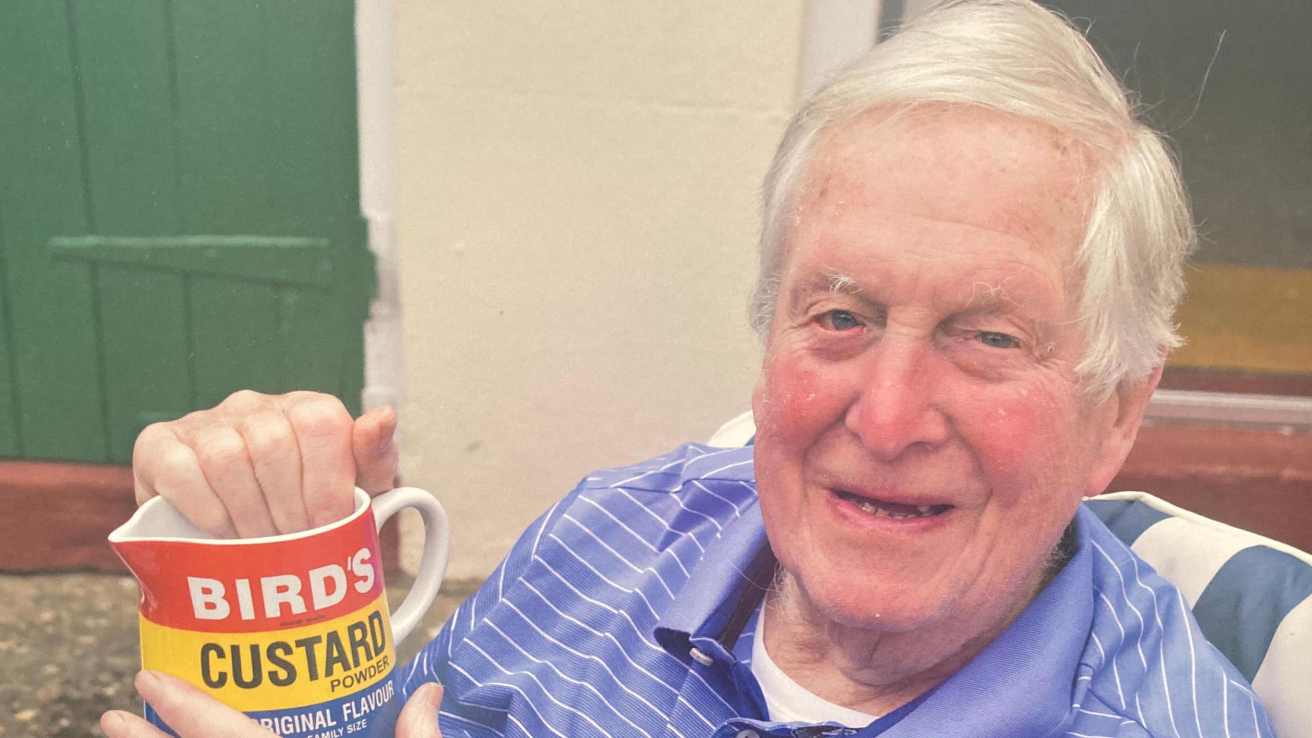Leslie aged 100, holding a Bird's Custard jug, in a chair, with short grey hair, and wearing a blue and white top. He is smiling, and a green door is behind him. 