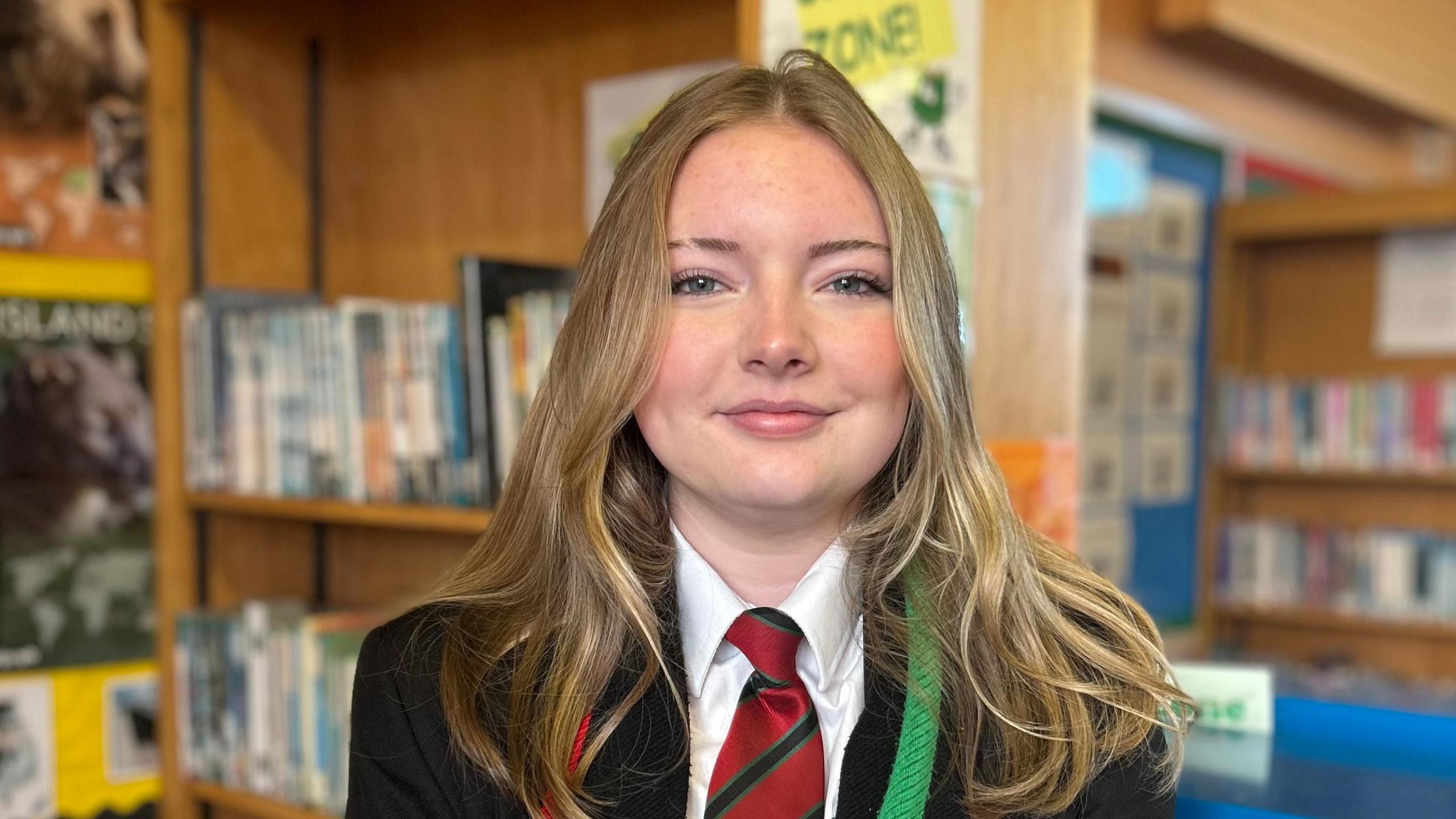 Lexi pictured in her school uniform, which is a black blazer with red and green trim, a white shirt and a red, light and dark green striped tie. She has long blonde hair and is standing in a library. 