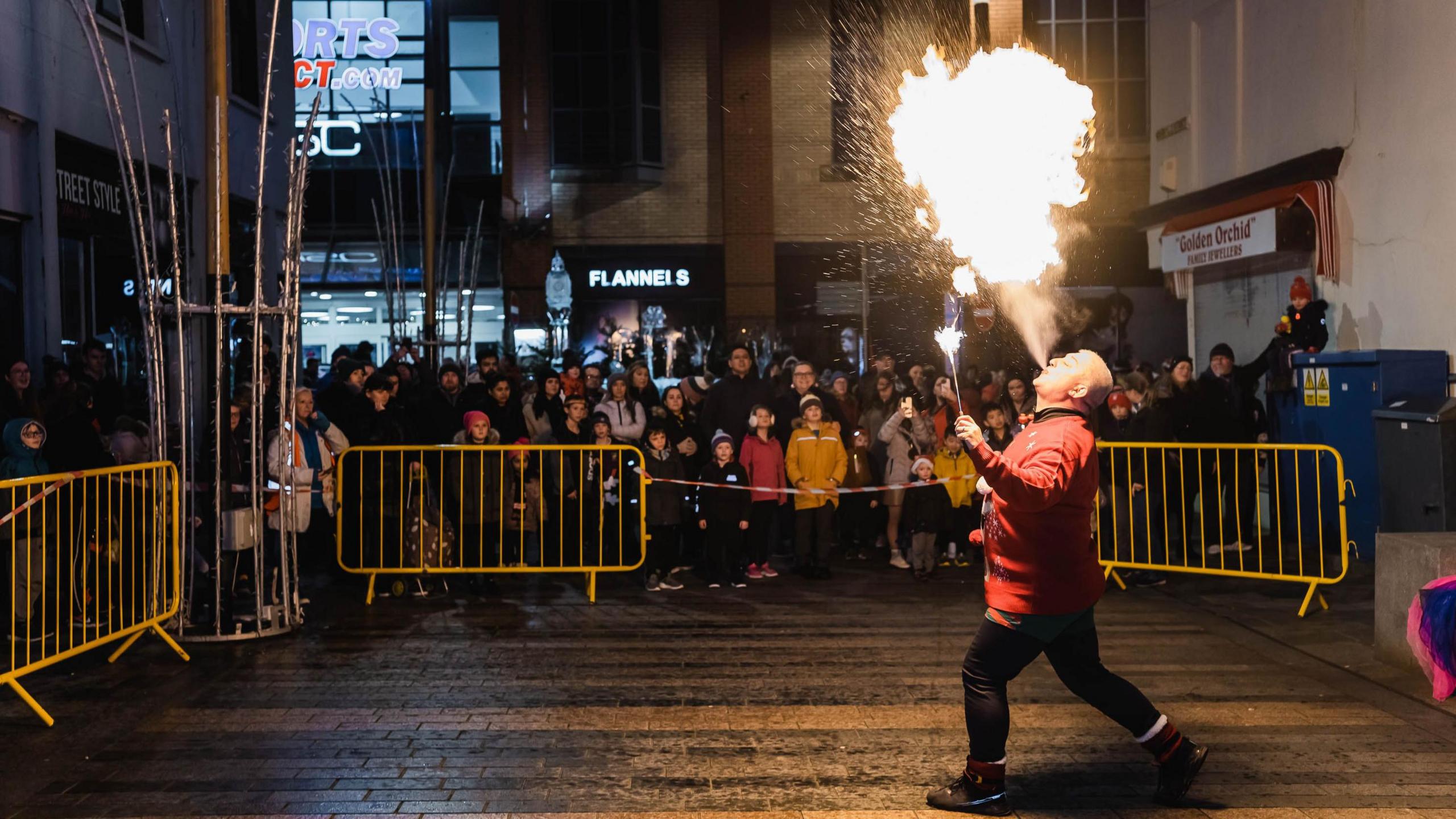 A firebreather blowing a plume of fire out of his mouth up into the air, in front of an audience of people in a town street at night. 