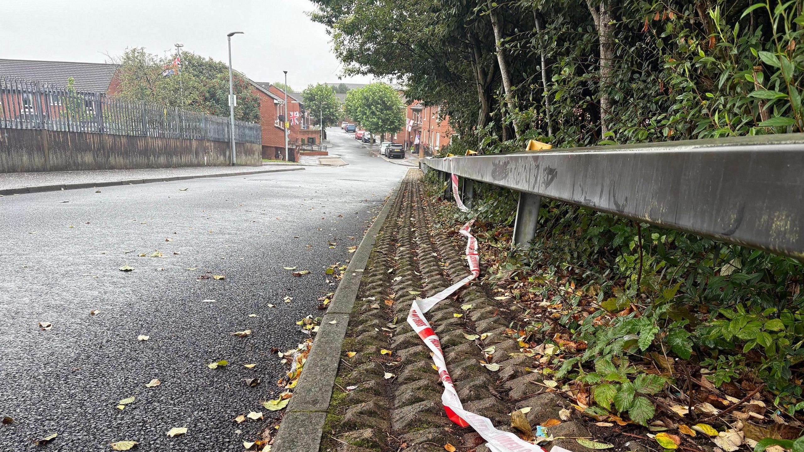 The remains of a police corden tape which had blocked off Bentham Drive in Belfast on Saturday. The red and white tape is lying near the kerb along a steel safety barrier. There are red brick houses in the distance and a wall with a metal fence runs alongside the other side of the road.
