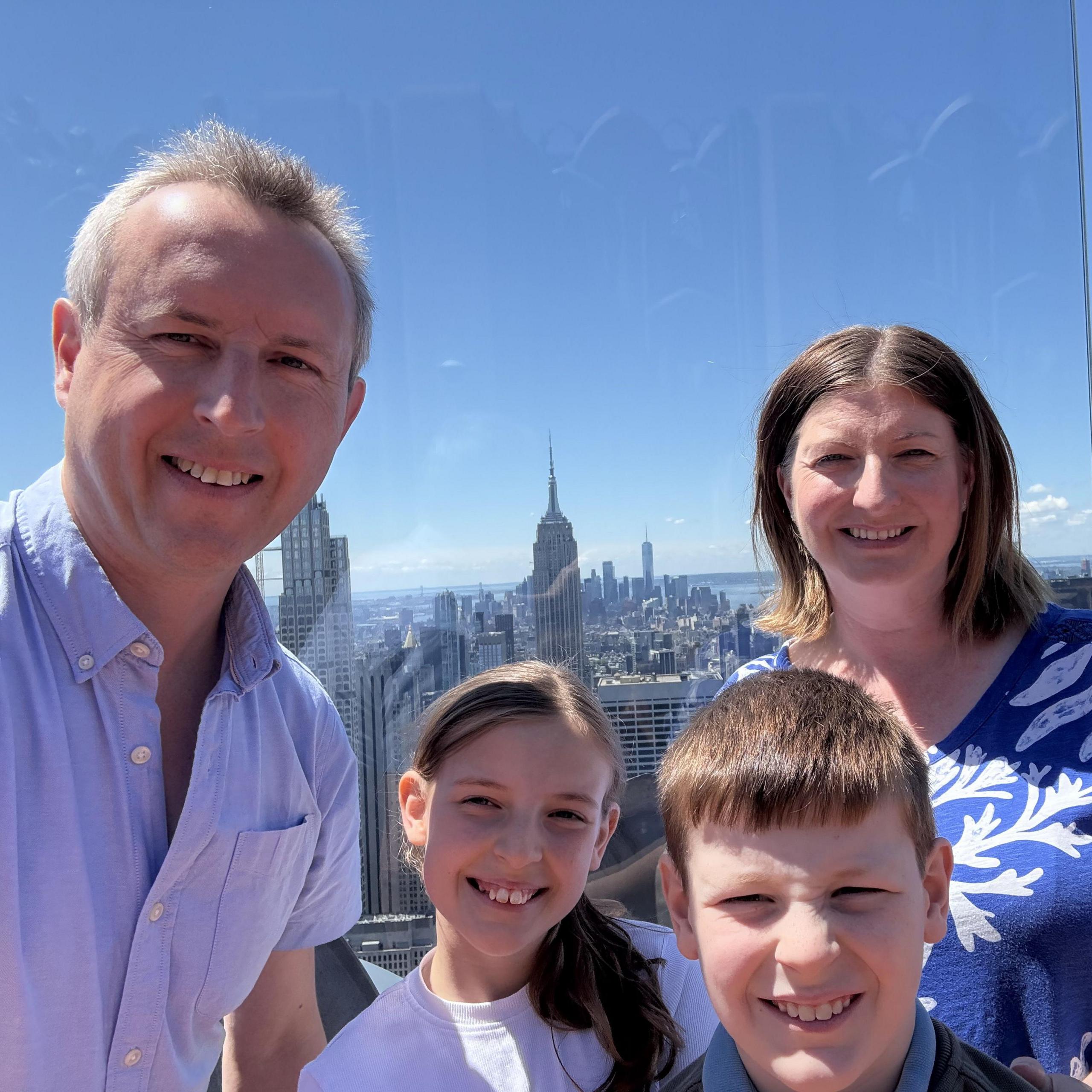 David Harris and his family take a selfie while posing on top of the Rockefeller Center in New York. The New York skyline and its skyscrapers can be seen in the background.