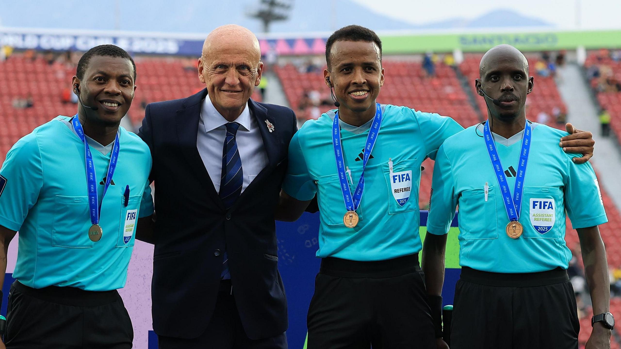 Pierluigi Collina, wearing a dark suit, blue striped tie and white shirt, is flanked by Omar Artan and two other officials who are all wearing light blue shirts and dark shorts. All three officials have medals around their necks on blue ribbons, and a sparsely populated stand is seen out of focus in the background