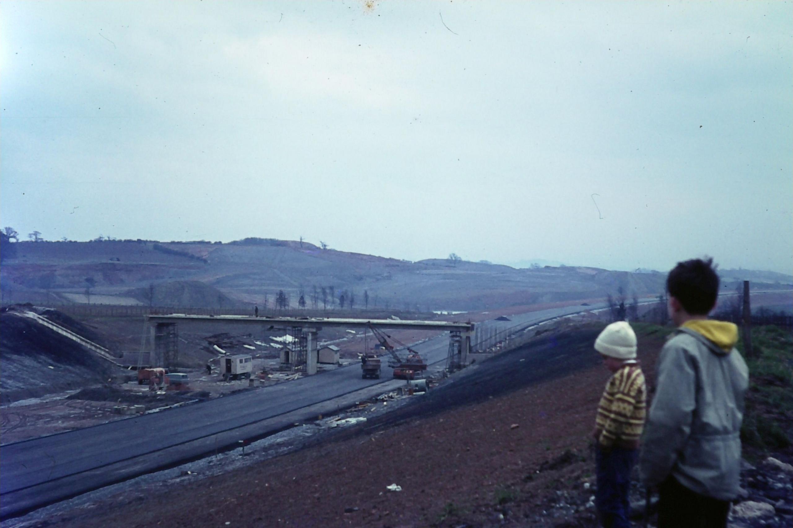 A vintage photograph with distinctive discolouration showing two children looking from an embankment over a motorway and bridge being built through the countryside