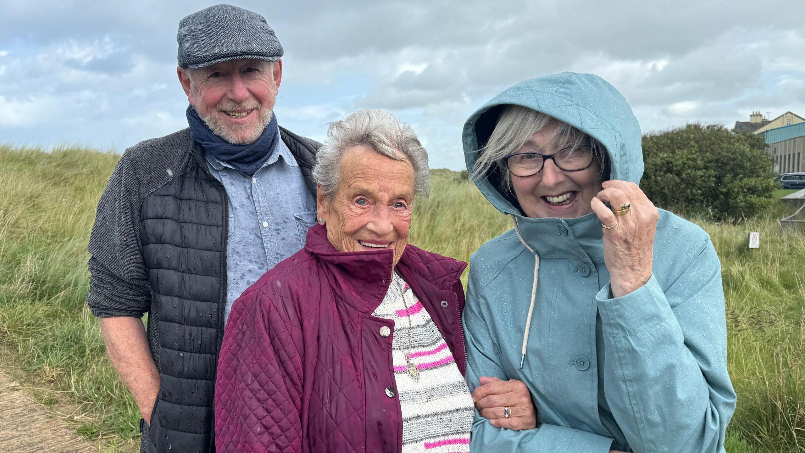 A man in a grey flat cap and jacket, a lady in a pink coat and another lady with glasses in a blue coat with the hood up