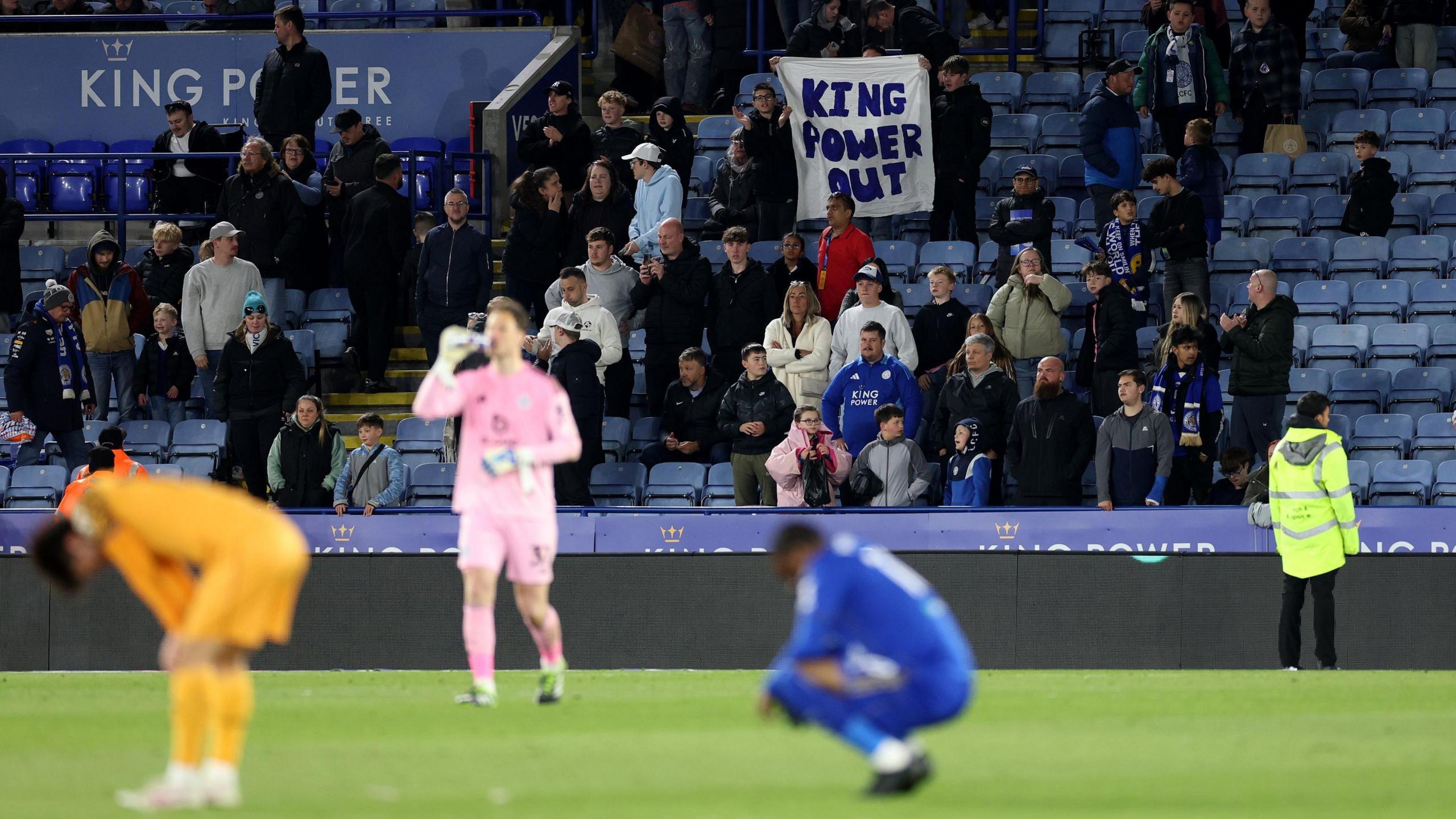Leicester City fans hold up a sign demanding the King Power Group sells the club, while in the blurred foreground a Foxes player can be seen hunched over in disappointment after relegation 