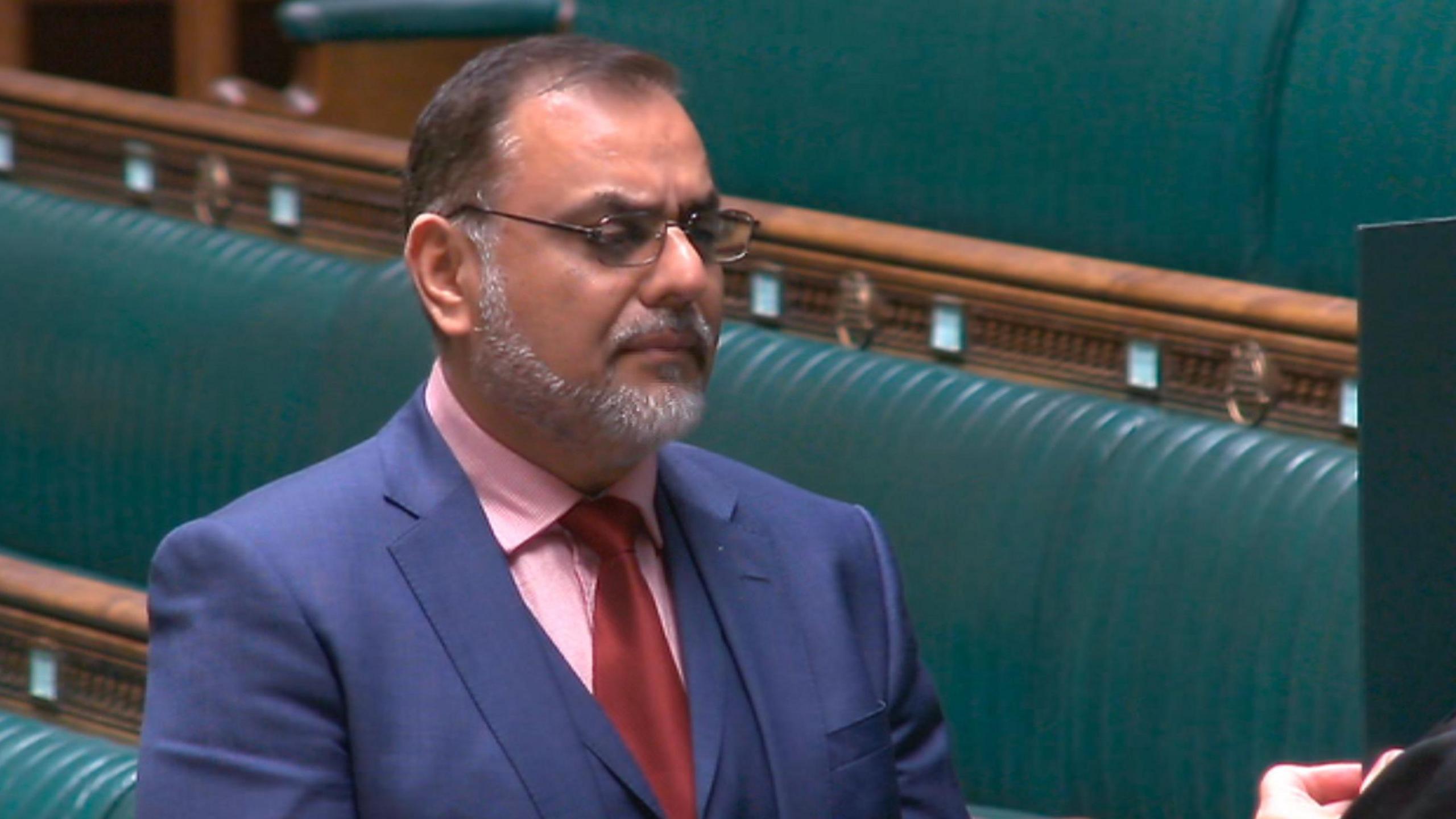 Ayoub Khan in a blue suit, pink shirt and red tie, being sworn in. The green benches of the Commons can be seen in the background.