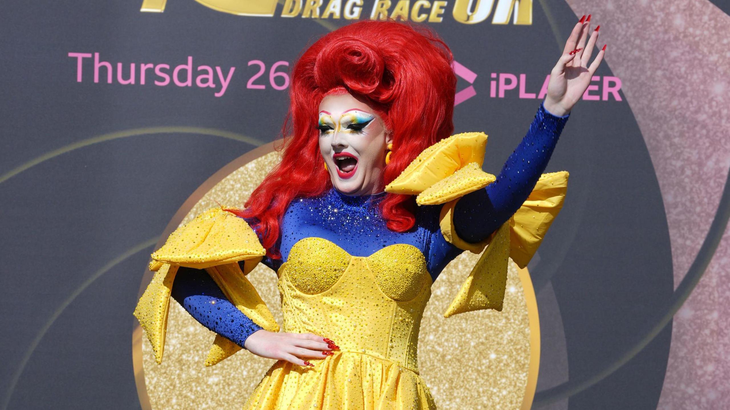 Drag queen Charra Tea wears a red wig, yellow bedazzled dress and long bedazzled blue sleeves.