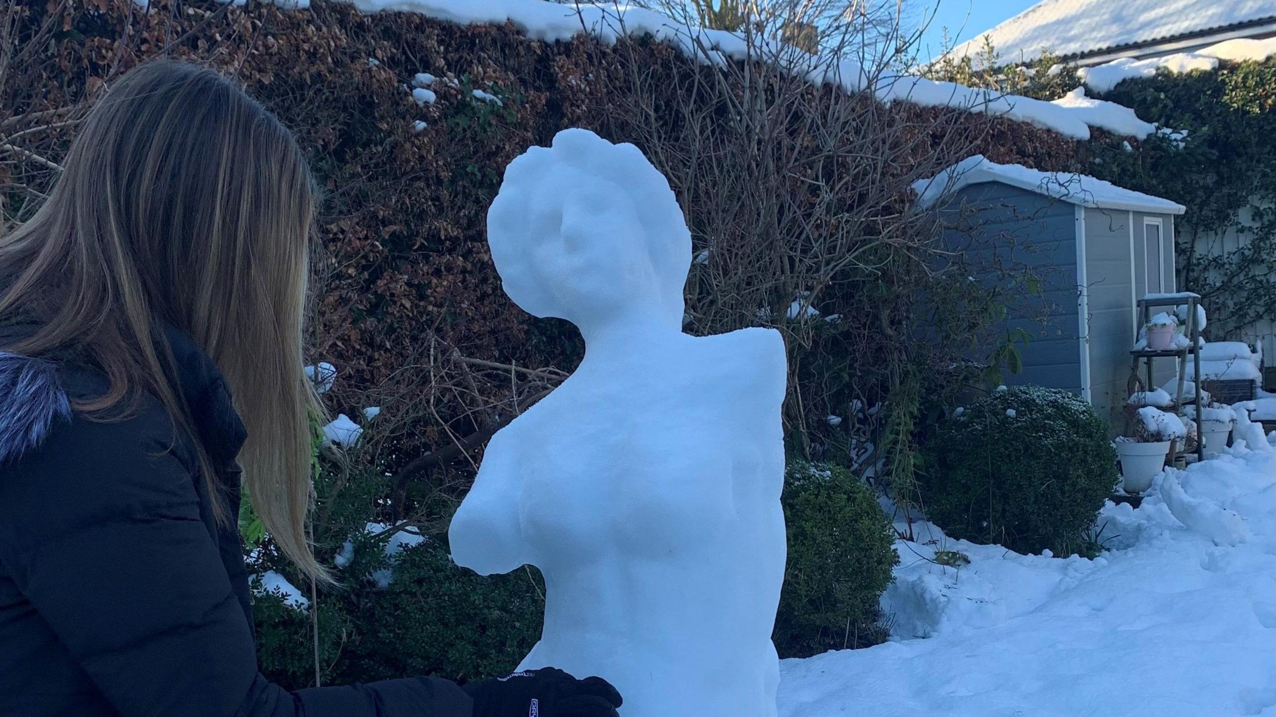 A teenage girl with long blonde hair in a black duffel coat builds a snow sculpture of the Venus de Milo.