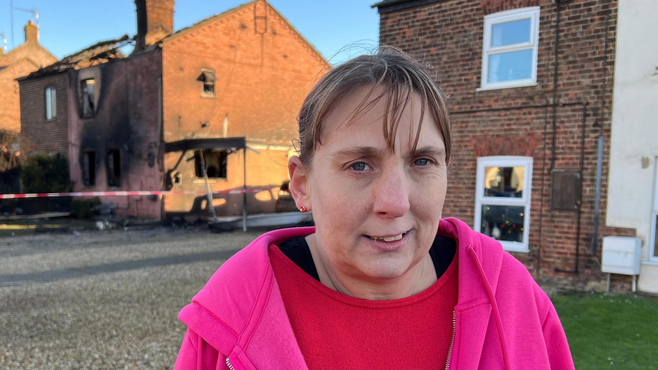 Karolyn has a blonde-brown hair tied back in a ponytail. She is wearing a pink hoodie over a red shirt. Underneath is a black base layer. She is standing in front of a burnt out house in the far distance on the left.