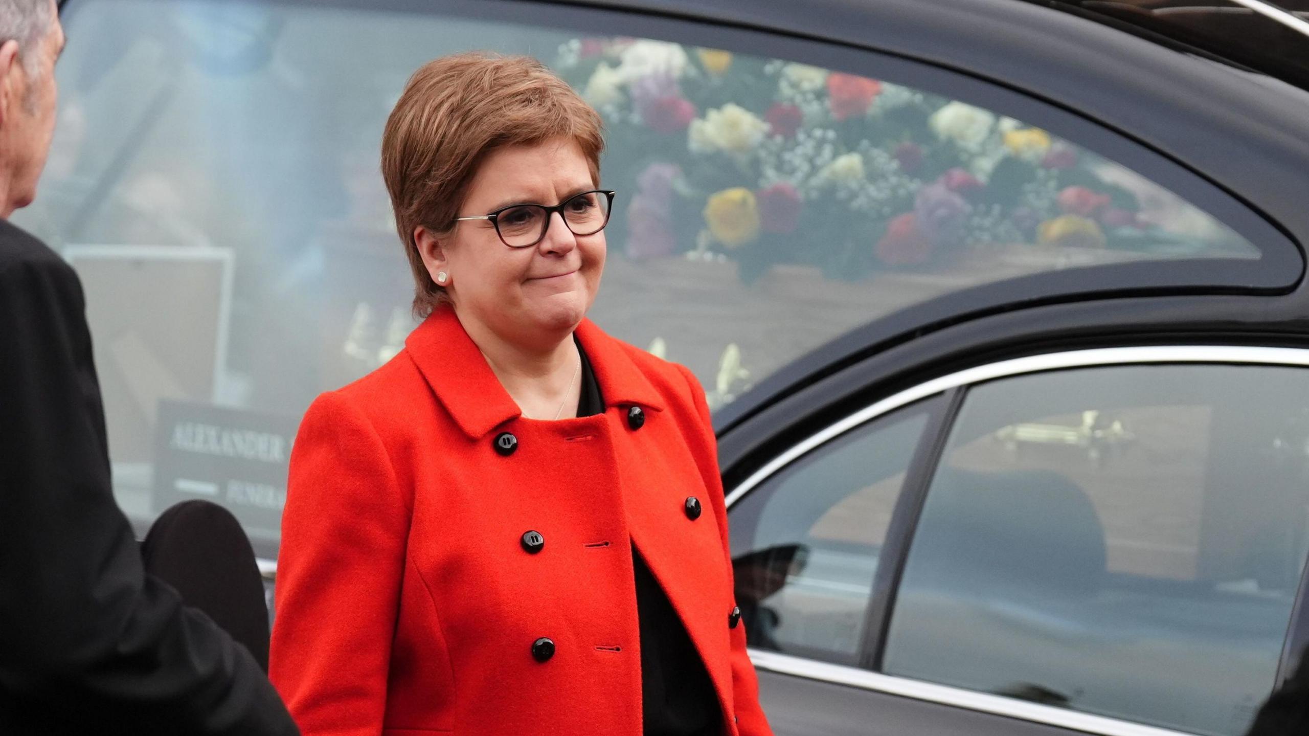 Sturgeon wearing a bright read coat stands in front of a hearse with a coffin covered in flowers.