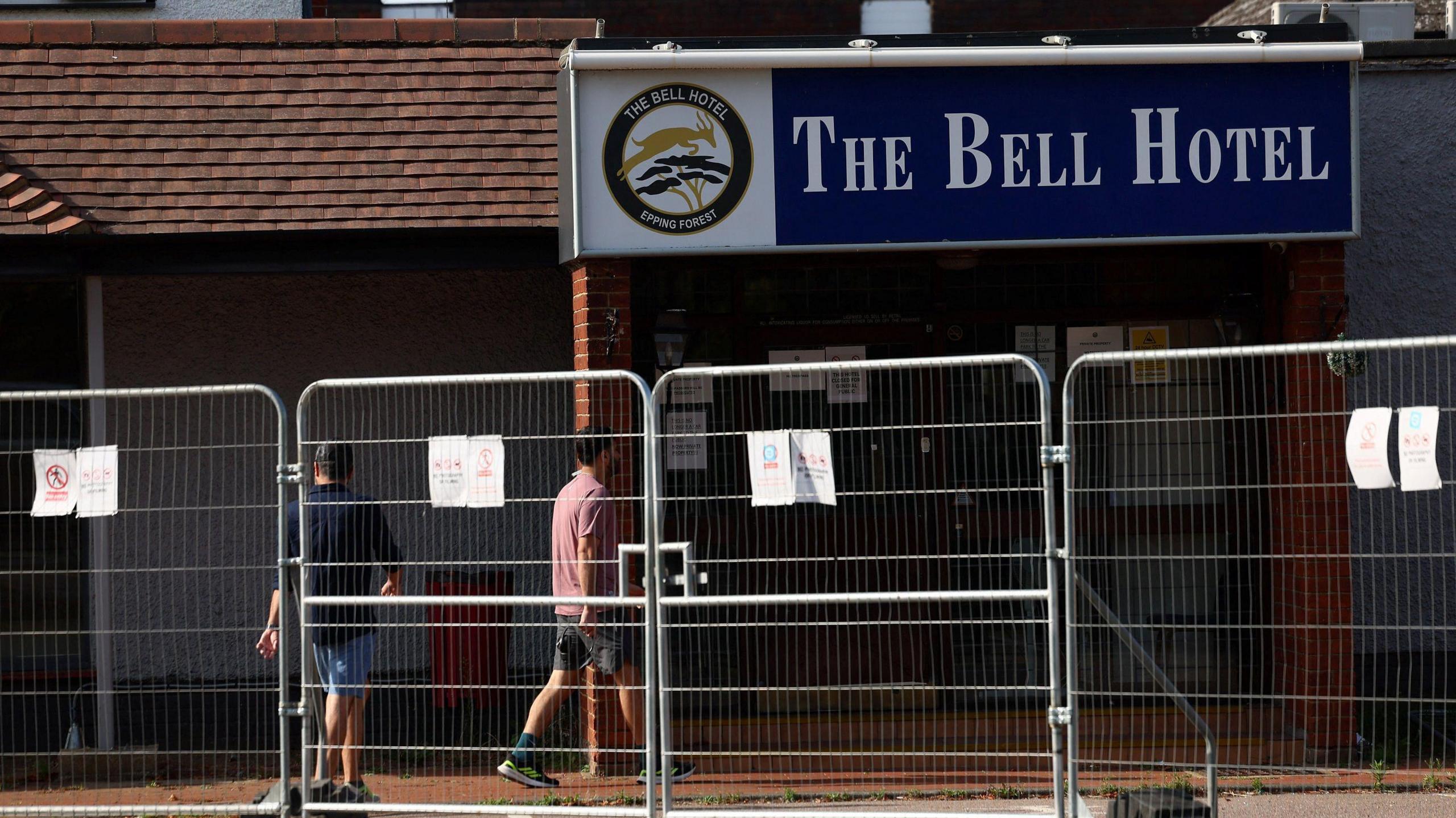 Two men can be seen walking into The Bell Hotel, which has a large sign above the door. The hotel is fenced off behind high metal temporary fencing. 