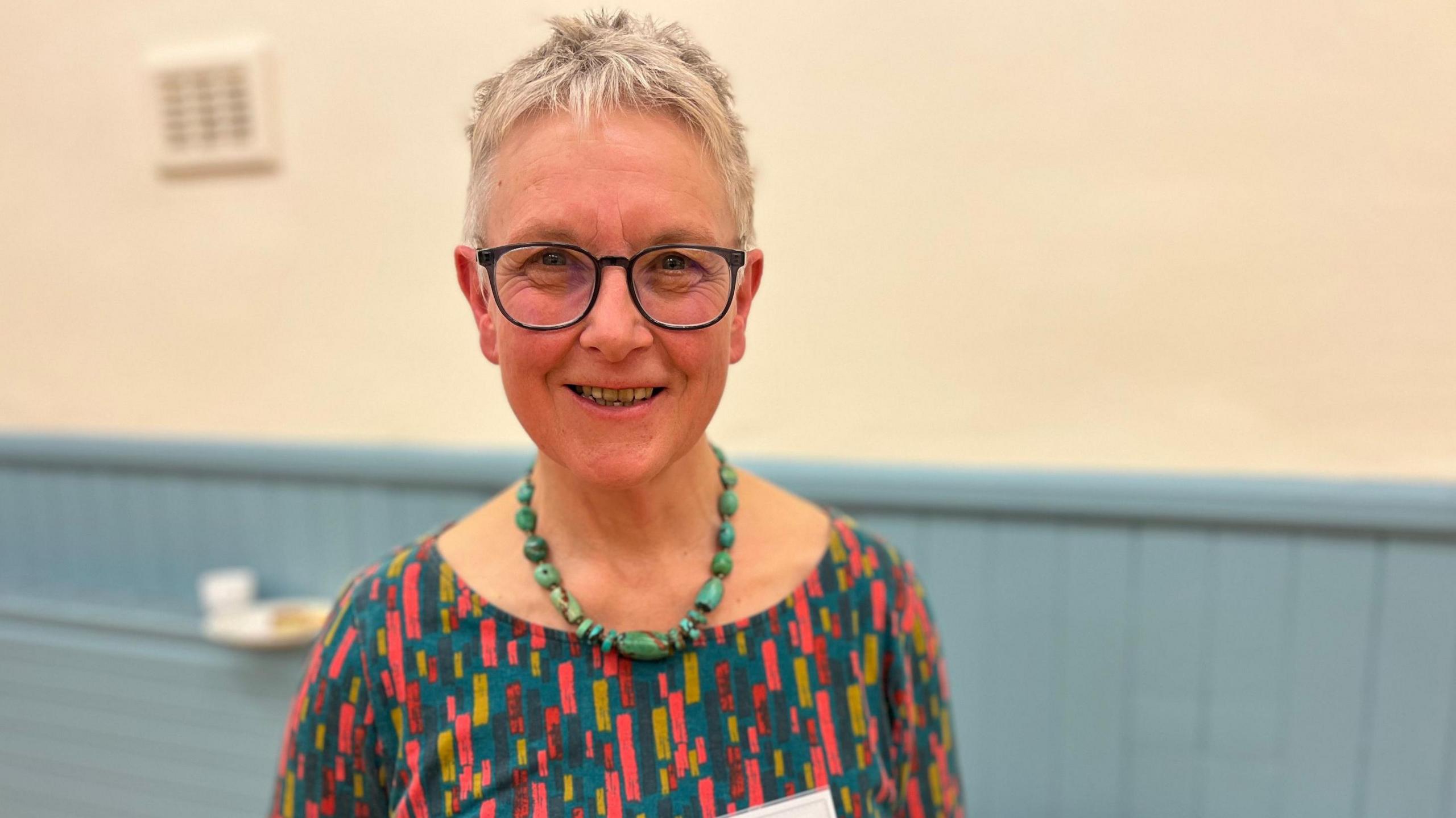 A woman with short, grey hair and glasses looks at the camera. She's wearing a multi-coloured top and a necklace of large green beads