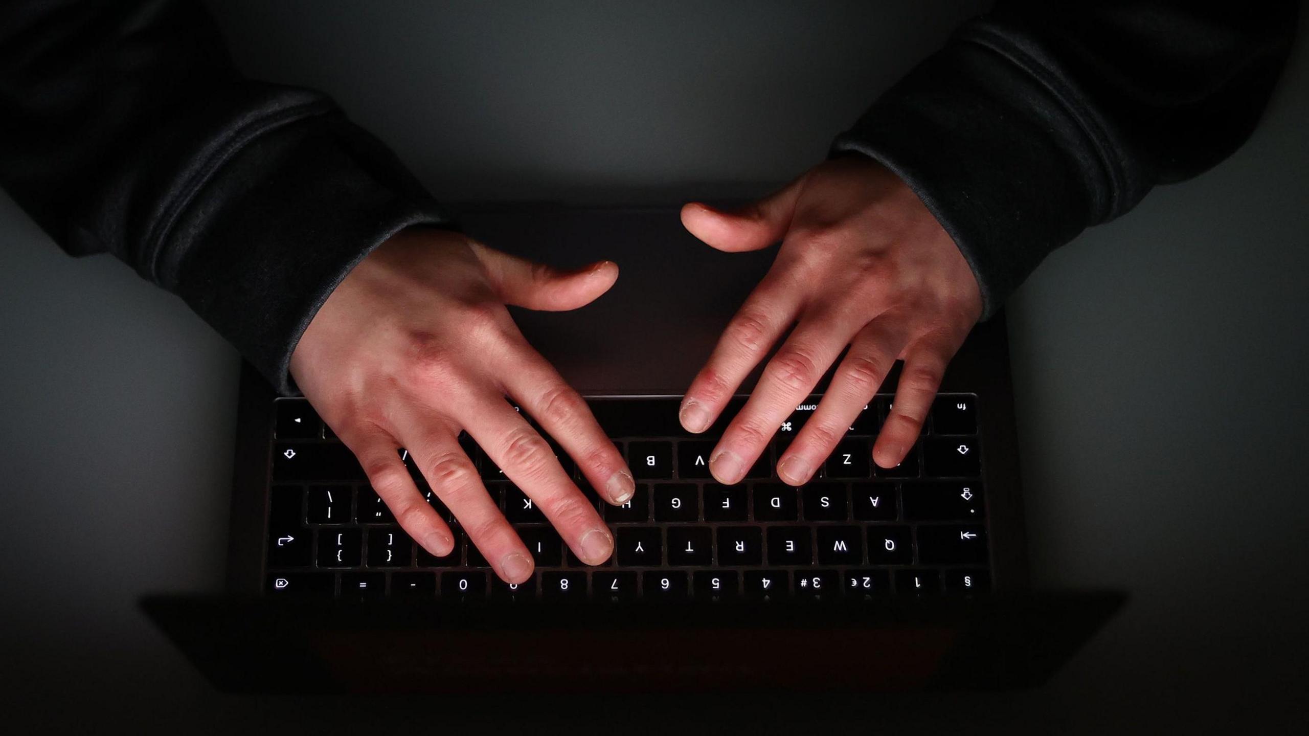 A birds-eye view angle of two hands typing on a black black keyboard, on a dark table surface.