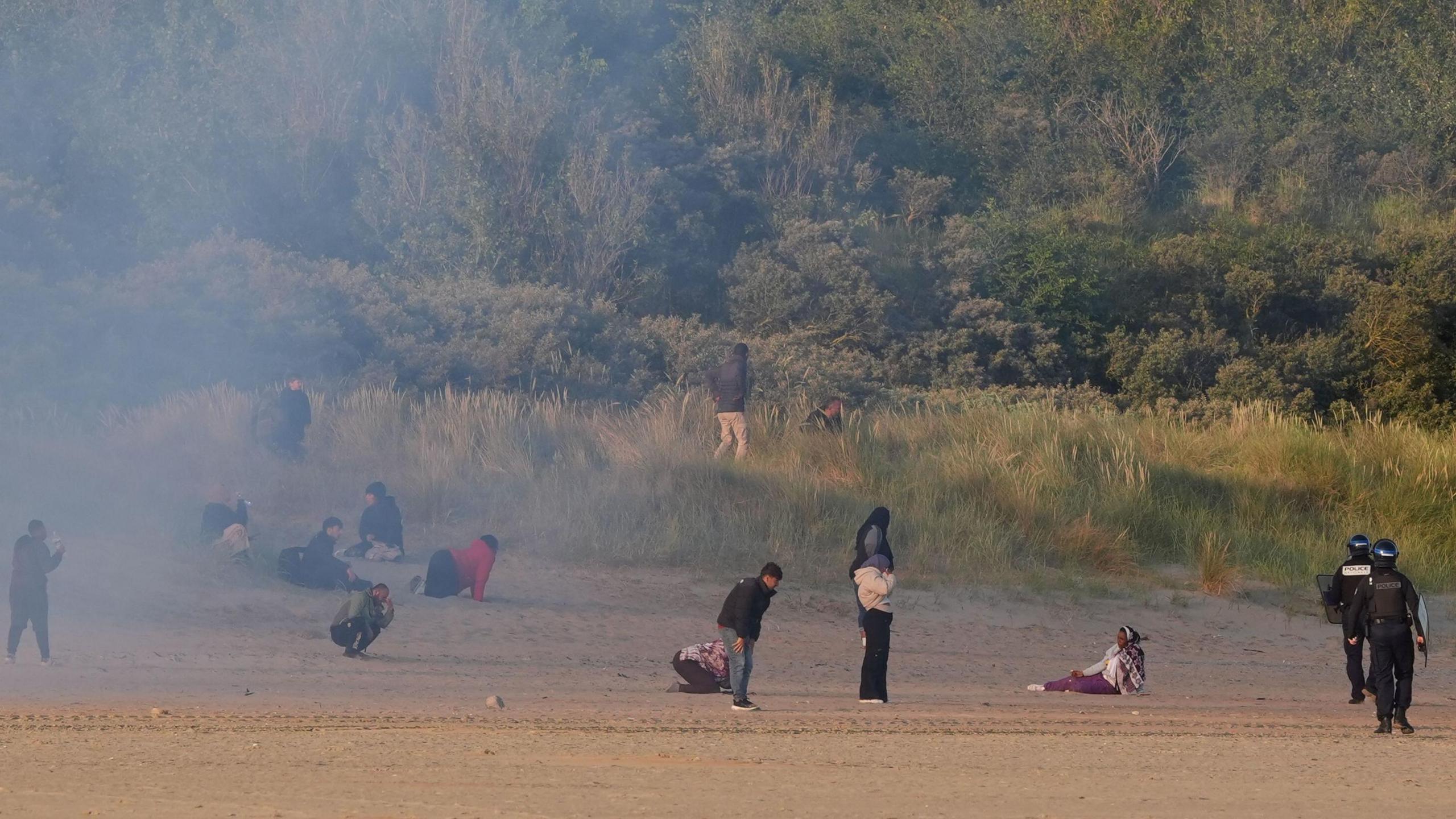 A disparate crowd of people stand in a cloud of gas, looking dejected, face Frenc police officers wearing riot gear near bushes along the treeline of a beach in Gravelines, France