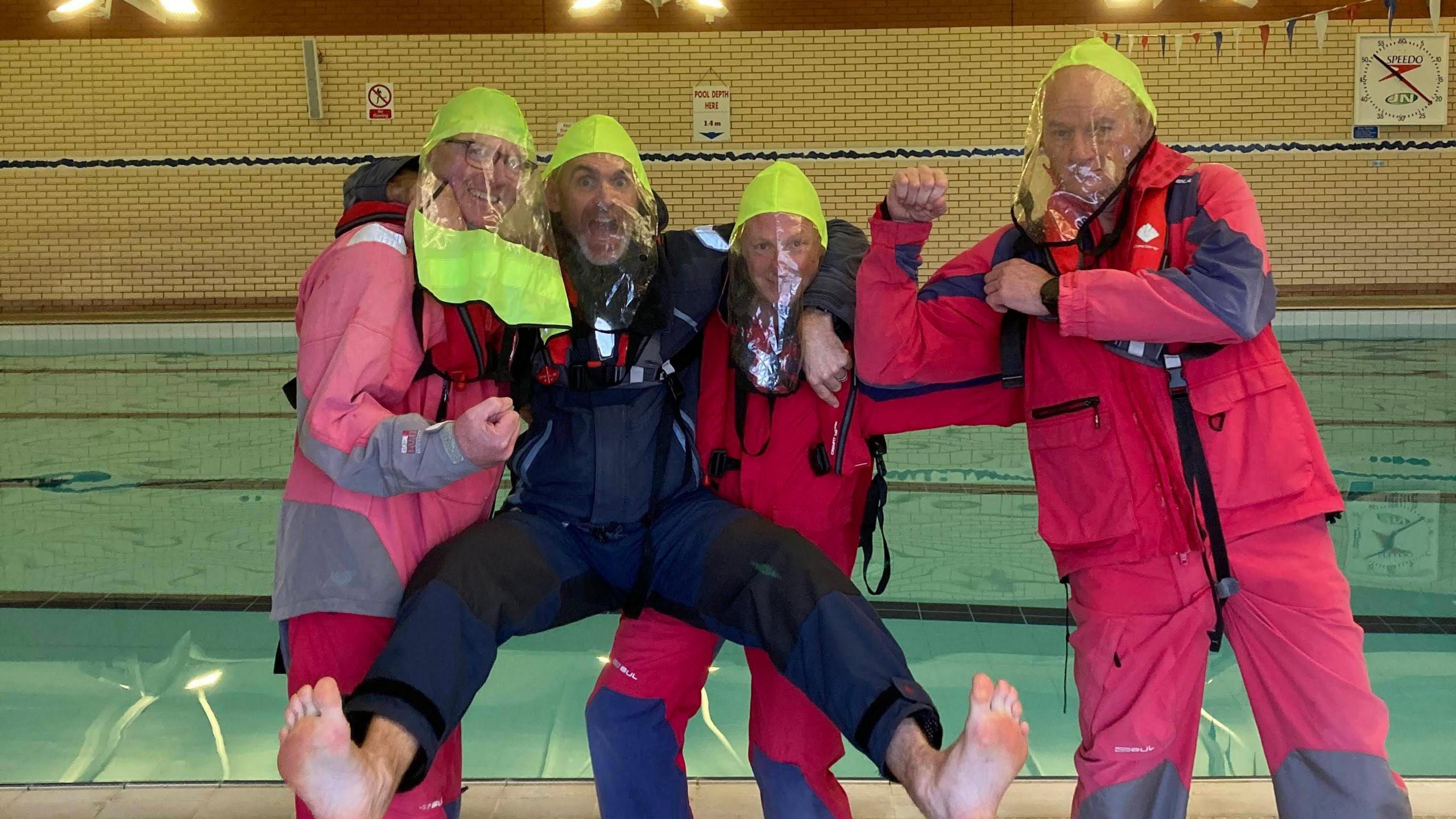 Four men in red and blue waterproof suits wearing yellow plastic hoods standing in front of a swimming pool and pulling silly poses. 