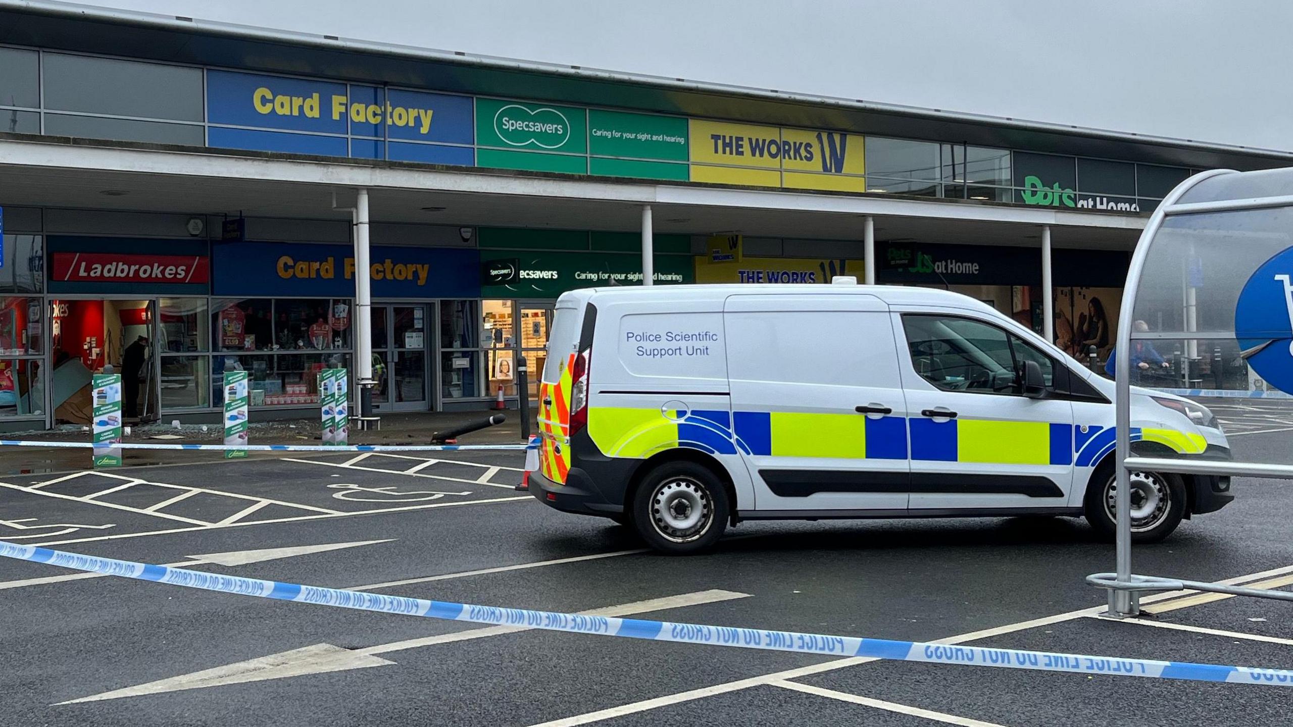 A white transit van with police scientific support unit on its back side panel and yellow and blue markings parked up in a car park behind blue and white police tape. Behind it is a single-storey row of shops under a low-lying roof, supported by pillars. Stepped back behind it are the shops, including a Ladbrokes with a destroyed door on its hinges, with its glass shattered on the ground. 