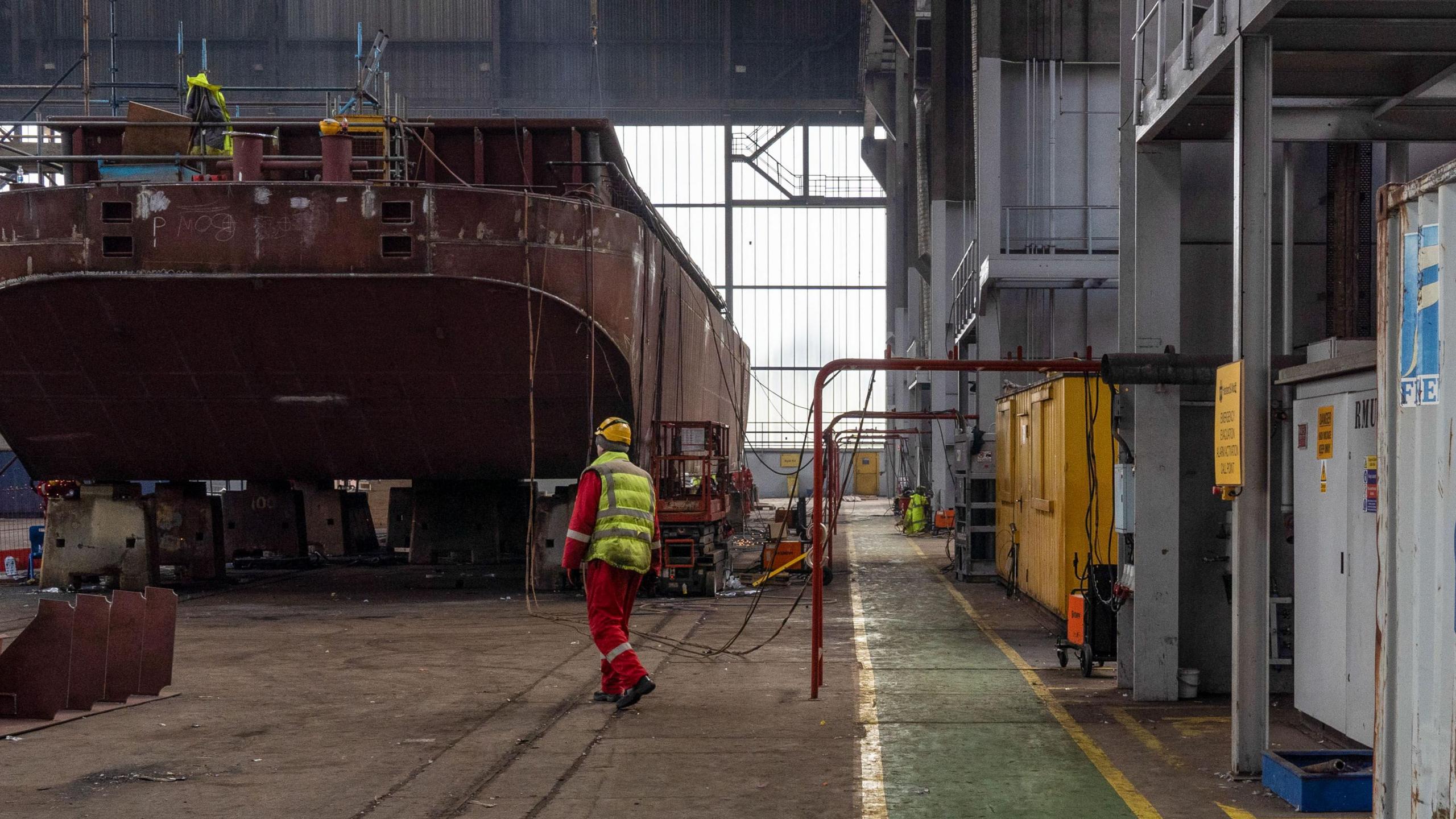 The production facility in Harland & Wolff Group Holdings shipyard in Belfast. A man is wearing a yellow hardhat, a high-vis jacket and a red jumpsuit.