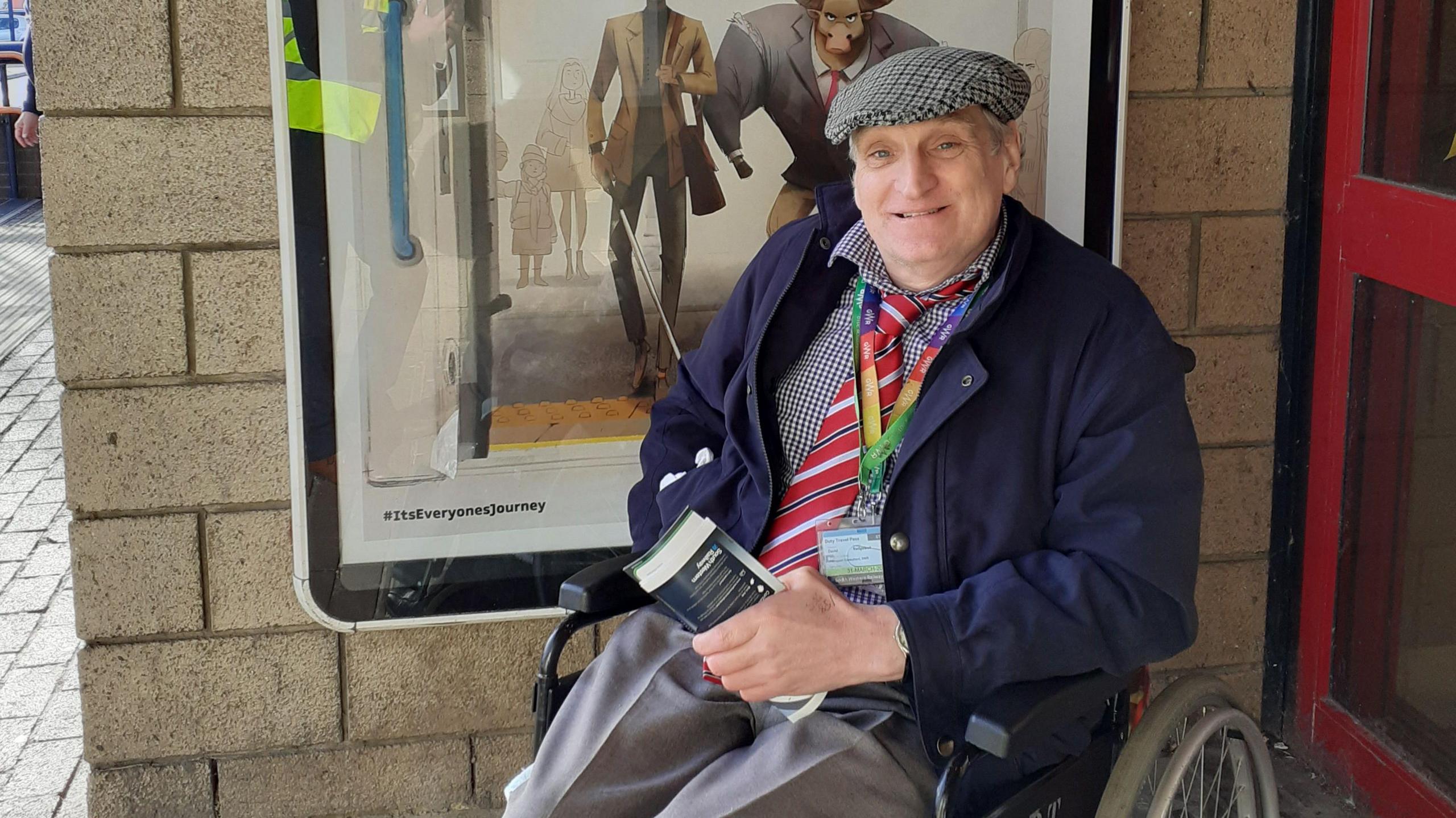 David Redgewell wearing grey trousers, a blue and white checked shirt and a navy blue raincoat and a checked cap. He is sitting outdoors in a wheelchair beside a door and a transport advertisement poster.