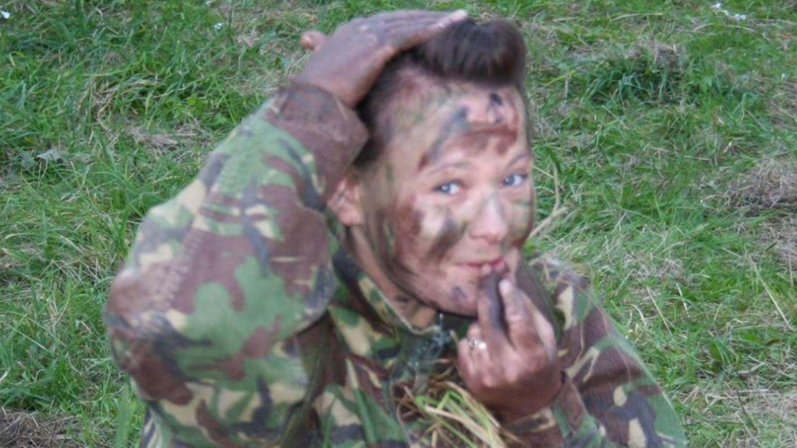 A woman in camouflage gear and combat body paint smiling while sitting on a grassy bank